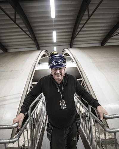 Sven Fahlén med hjälm och glasögon står på en metallgång i en industriell miljö med stora välvda strukturer i bakgrunden.