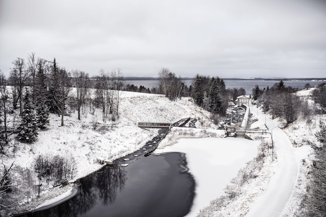 Snötäckt landskap med en flod, träd och en liten damm. En väg löper längs den frusna vattnet och himlen är täckt av moln.