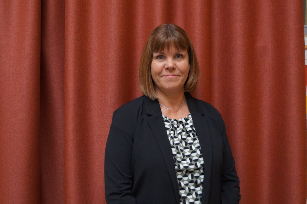 Lena Dalstam med axellångt hår, klädd i en svart blazer och mönstrad blus, står framför ett rött draperi.