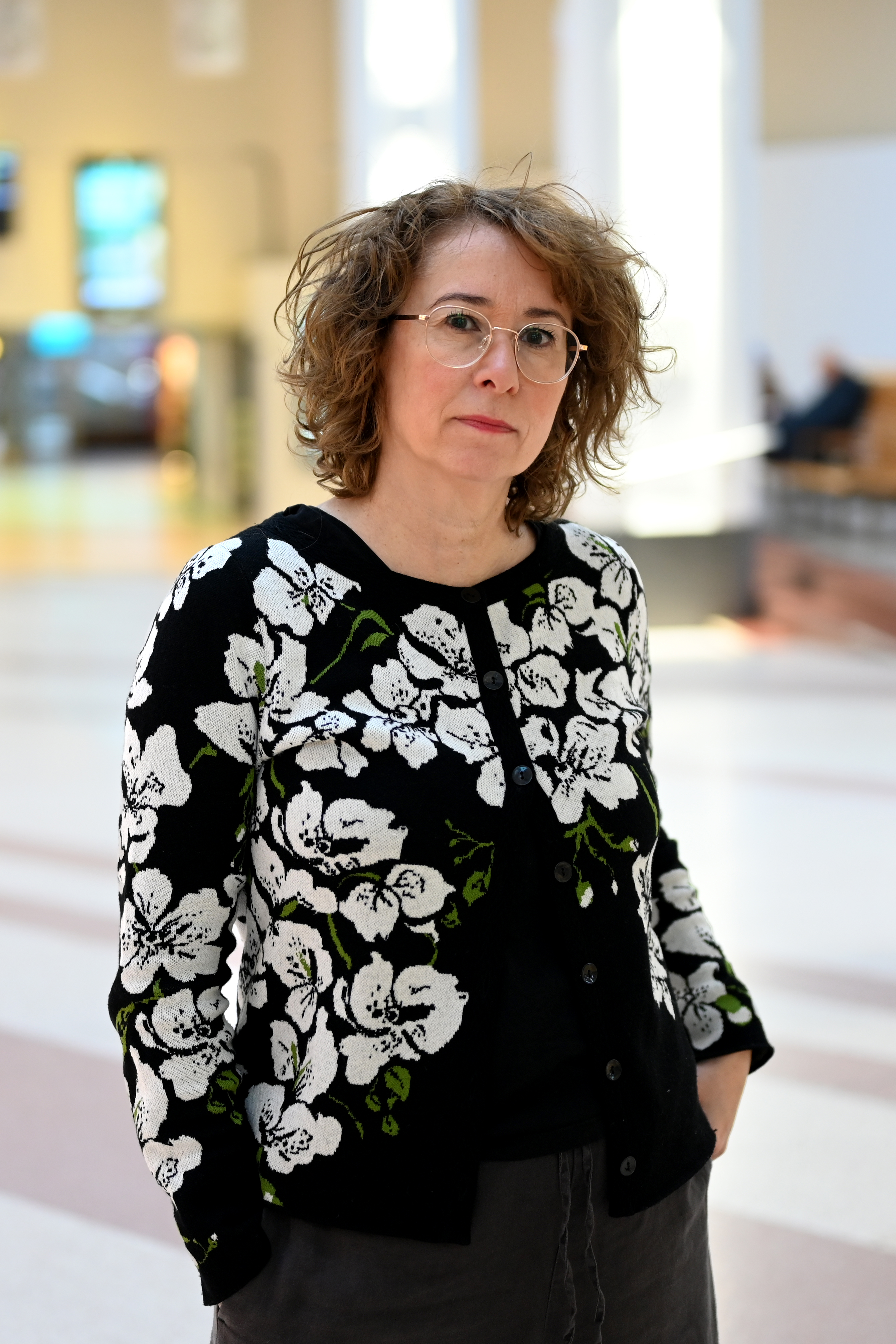 En kvinna med glasögon och en blommönstrad tröja står inomhus med händerna i fickorna, vänd mot kameran.