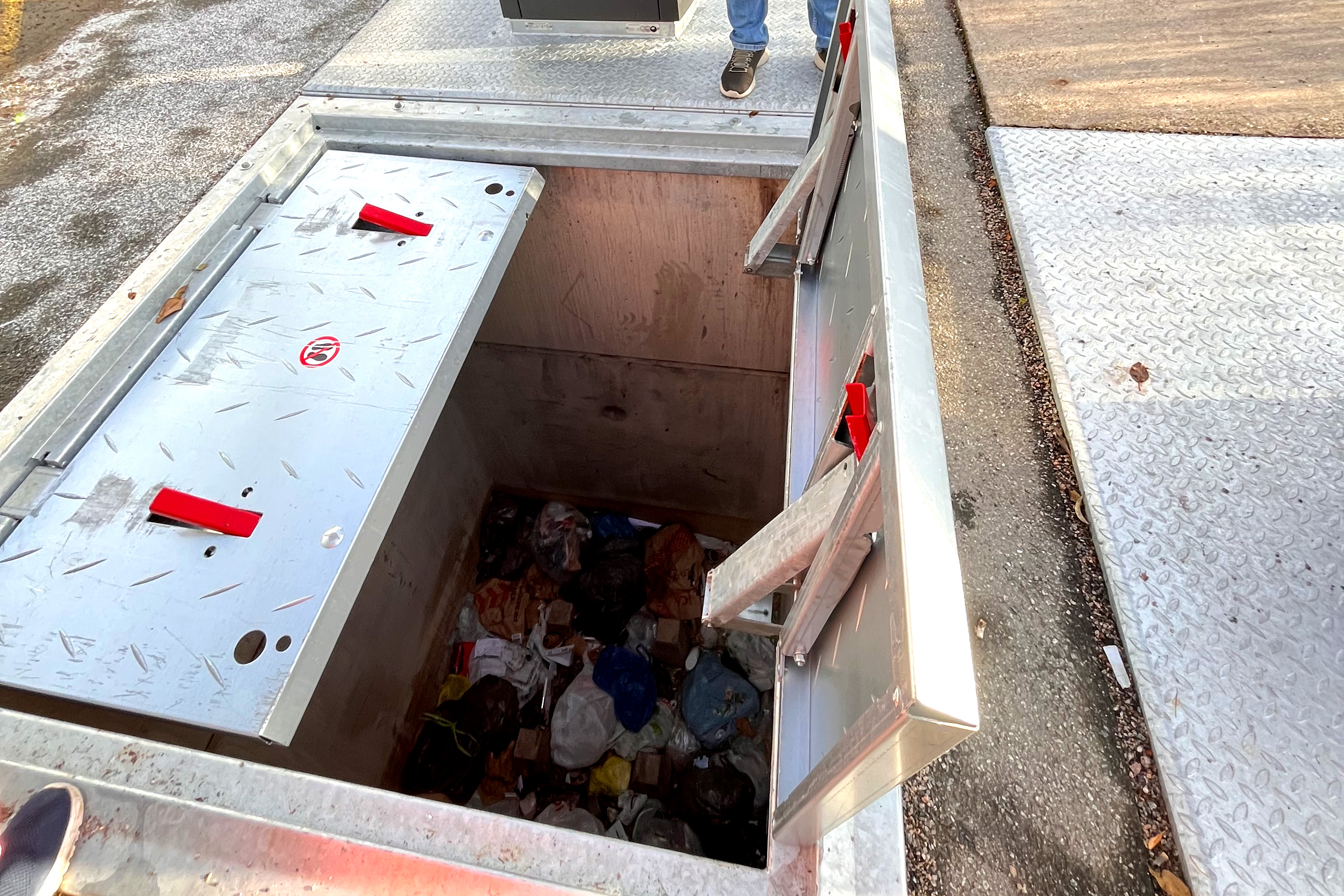 En stor metallcontainer med öppna lock är delvis fylld med olika sopor. En persons ben syns nära överkanten.