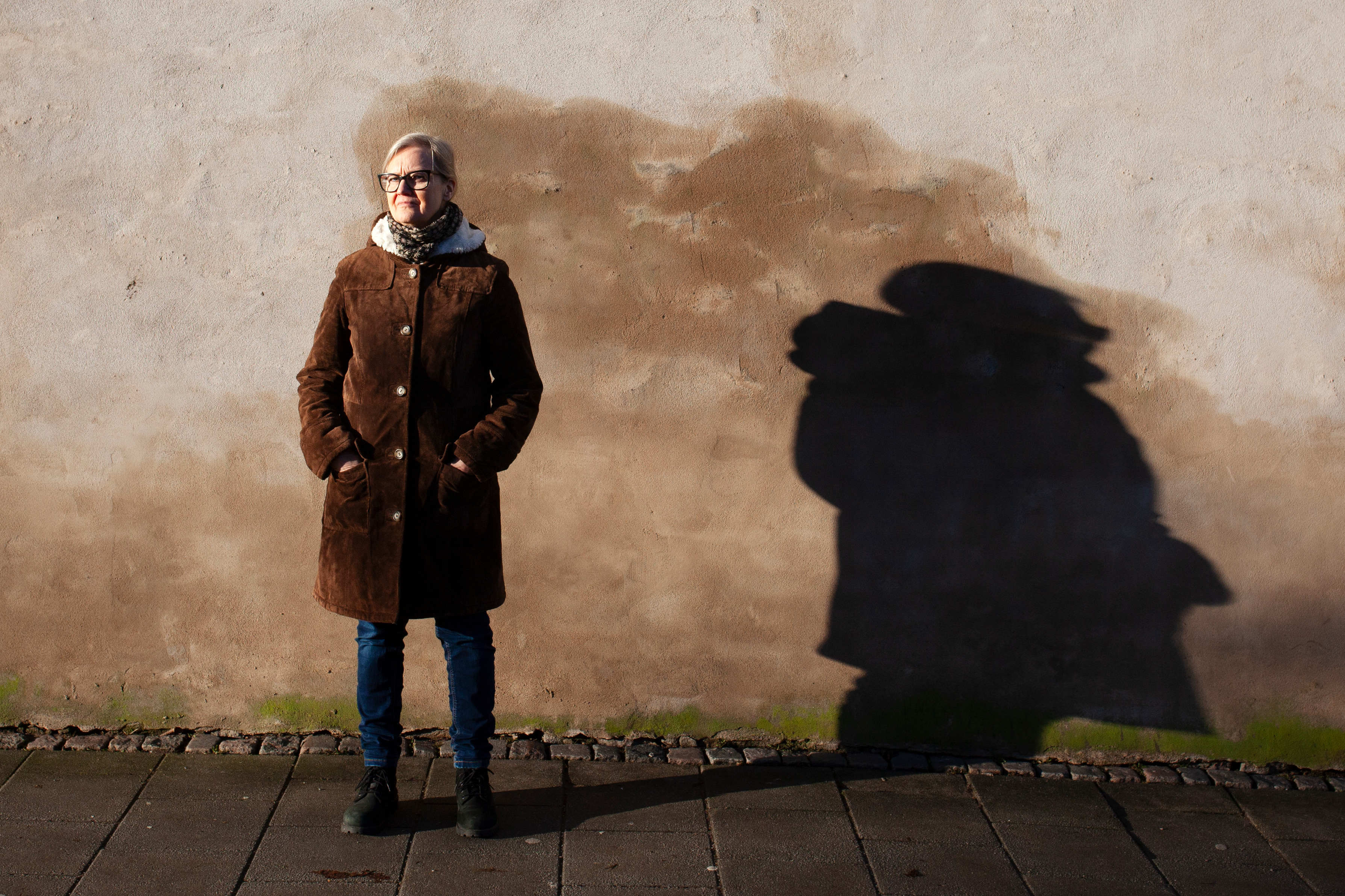 En person i brun rock står mot en solbelyst vägg med textur, vilket ger en tydlig skugga.