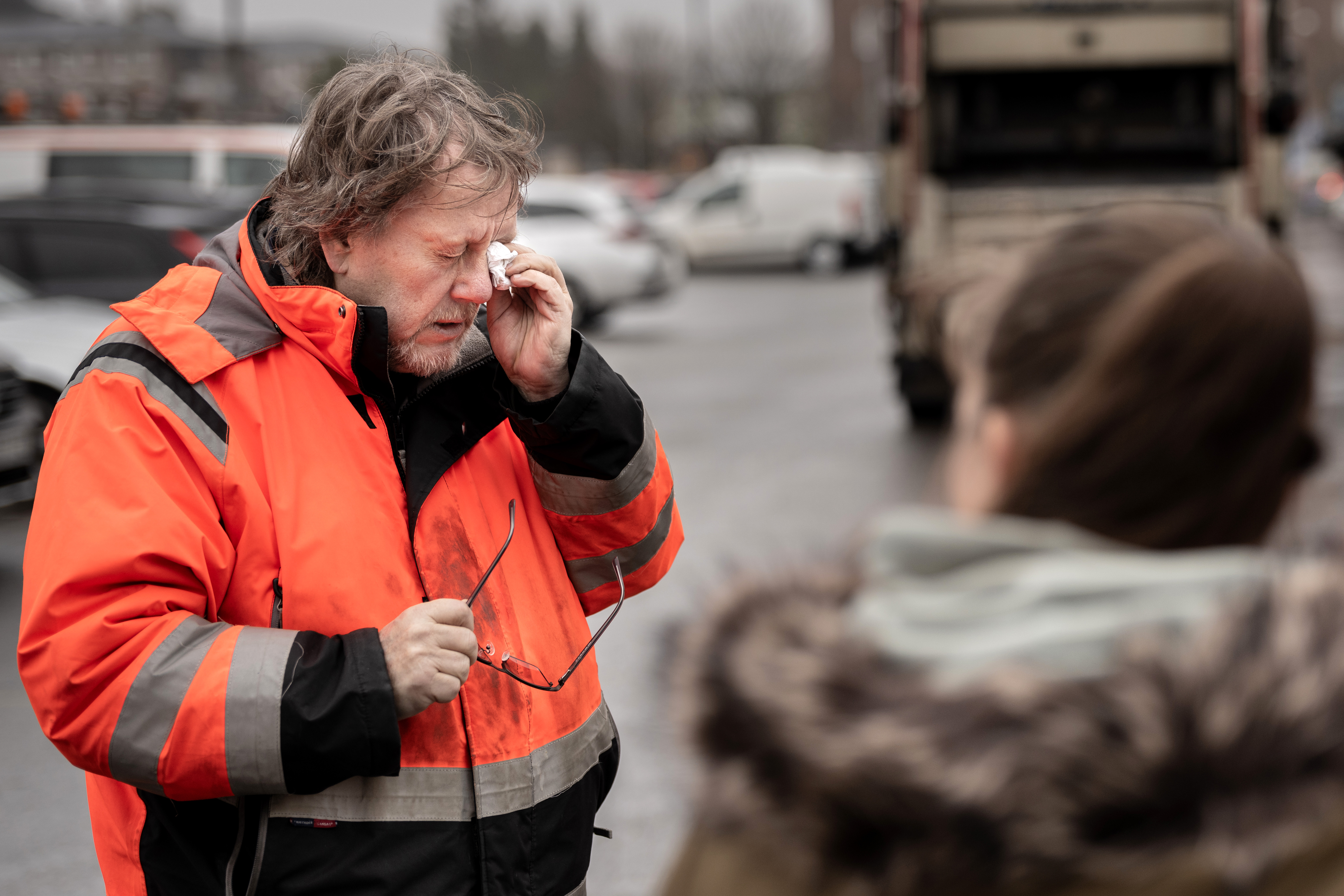 En man i orange jacka torkar sitt öga med en näsduk och håller ett par glasögon i handen. Han står på en parkeringsplats, och det finns en person i förgrunden.