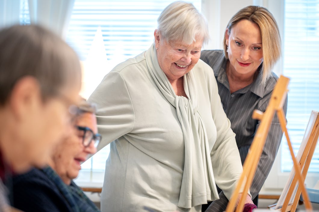 En grupp äldre vuxna deltar i en konstklass. Två kvinnor sitter och målar vid ett bord, medan en tredje kvinna står upp och ler.