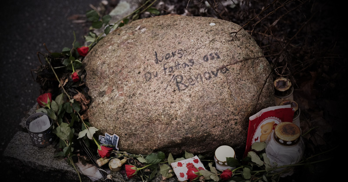 En sten med inskription är omgiven av röda rosor, ljus och olika små föremål, som tillsammans utgör ett minnesmärke.