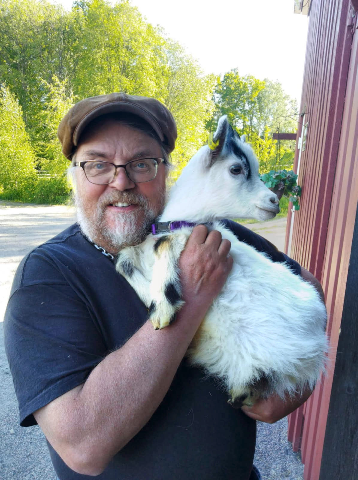 En man med keps och glasögon håller i en liten get utomhus nära en träbyggnad.