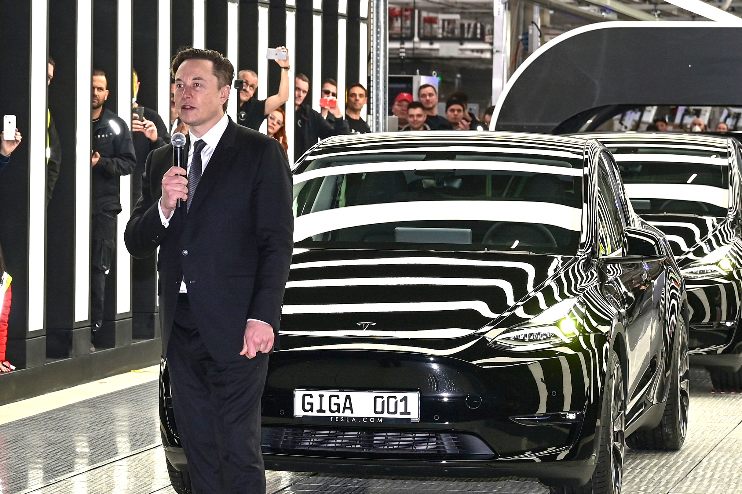 En person i kostym håller en mikrofon framför en svart bil märkt "GIGA G01" inne i en väl upplyst industrilokal med människor i bakgrunden som tar foton.