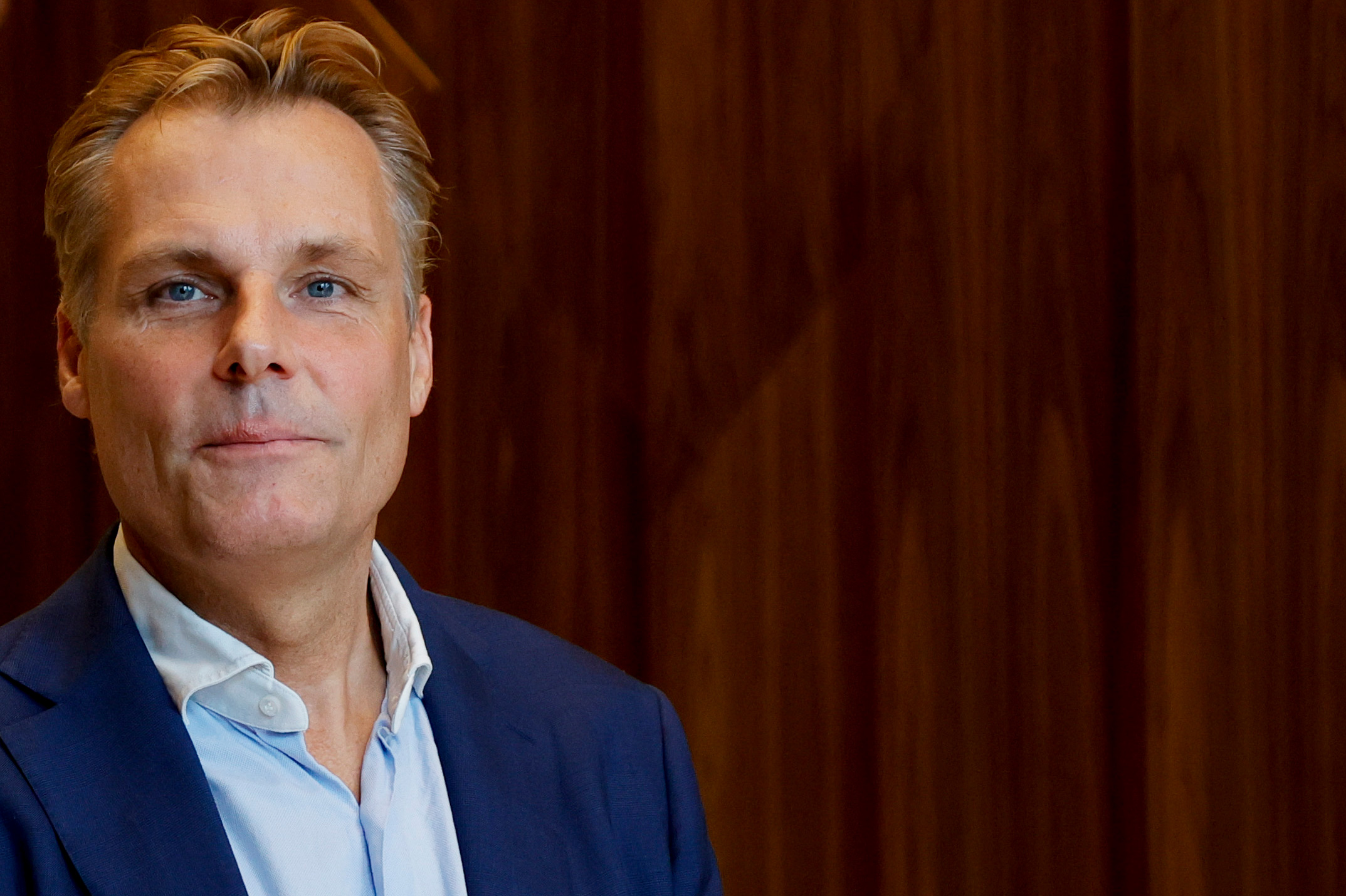 En person i blå kostym och vit skjorta står framför en trävägg.