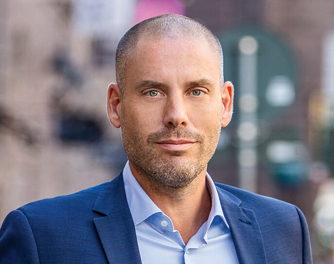 En man med rakat huvud och skägg står utomhus, klädd i en blå kostym och en ljusblå skjorta. Han ser direkt in i kameran, mot en suddig stadsbakgrund.