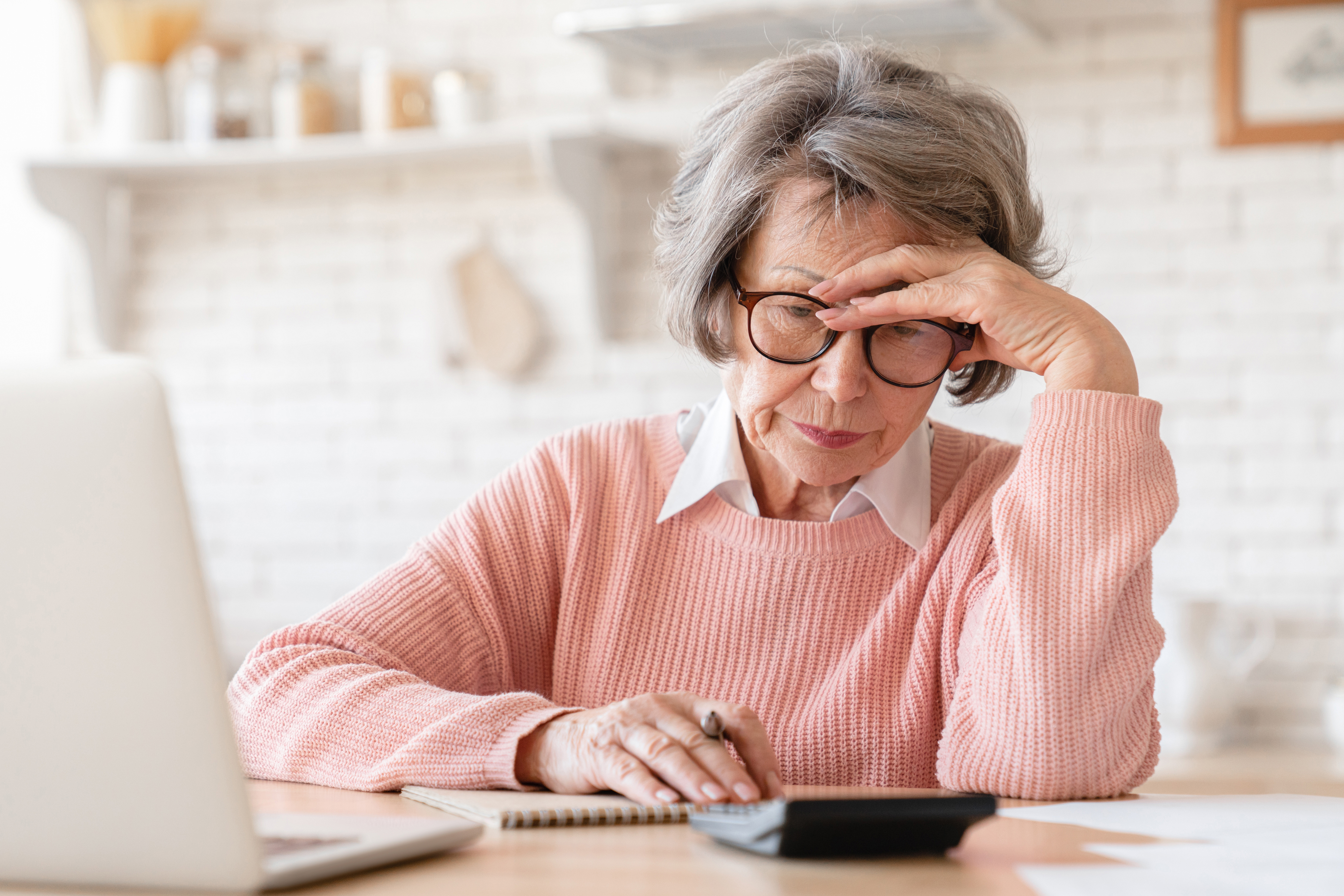 En äldre kvinna med glasögon och rosa tröja sitter vid ett bord med en miniräknare och en anteckningsbok framför sig.