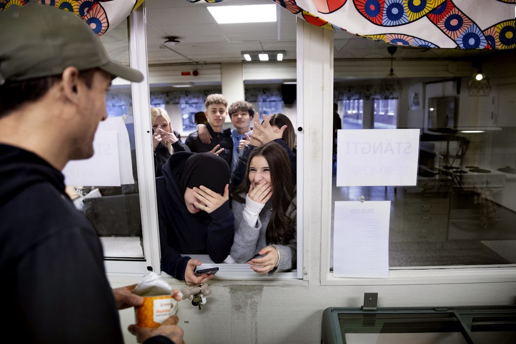 En grupp människor samlas vid en fönsterdisk. Vissa av dem håller händerna för ansiktena medan de tittar på René som håller i en behållare. Det finns skyltar synliga på glaset.