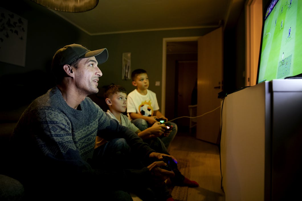 Tre personer sitter i ett svagt upplyst rum och spelar videospel på en stor skärm. Två barn håller i kontroller medan en vuxen tittar på, och alla är koncentrerade på spelet.