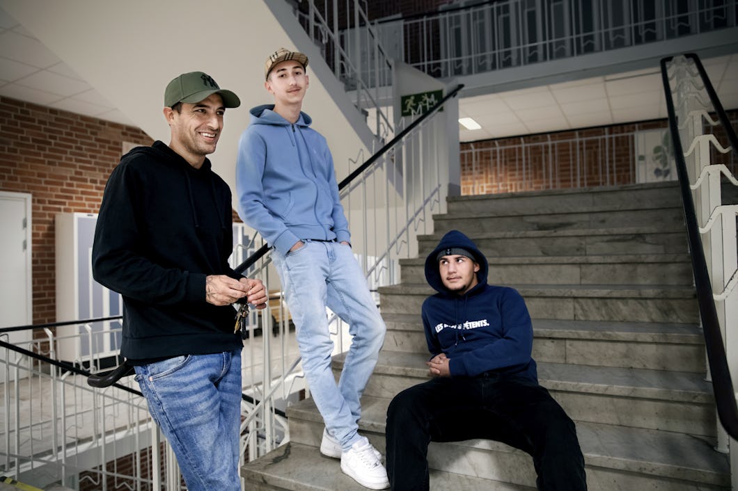Tre personer i vardagskläder är på en trappa. Två står och en sitter. I bakgrunden syns tegelväggar och metalräcken, vilket antyder att platsen är inomhus.