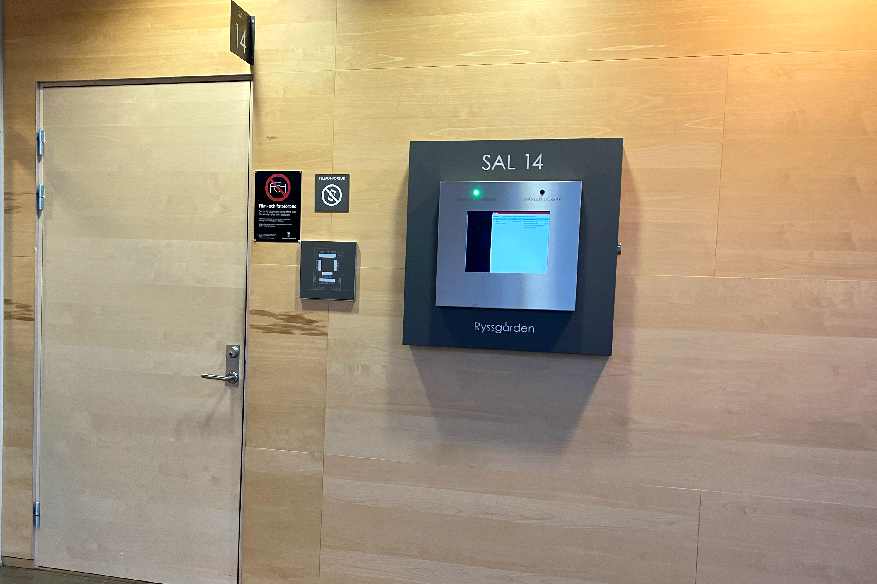 Trävägg med en dörr och en elektronisk skärm märkt "Sal 14." Rökförbudsskylt och handspritdispenser i närheten.