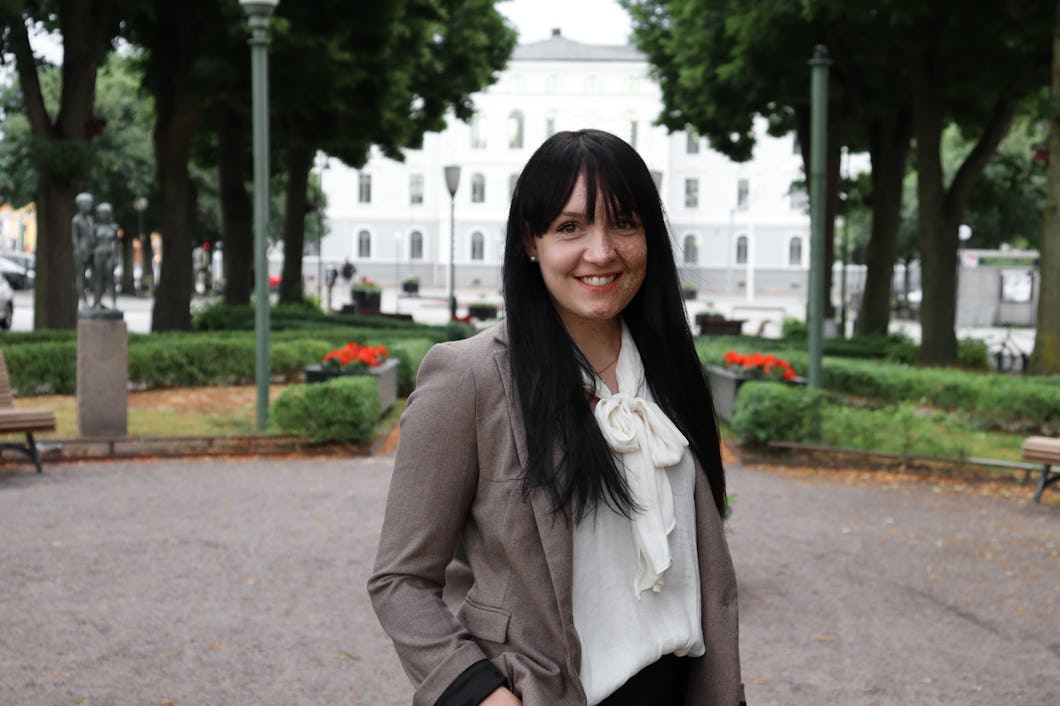 Sara Swahnström med långt mörkt hår står i en park och ler. Hon har på sig en beige blazer och en vit blus. I bakgrunden syns träd och en byggnad.