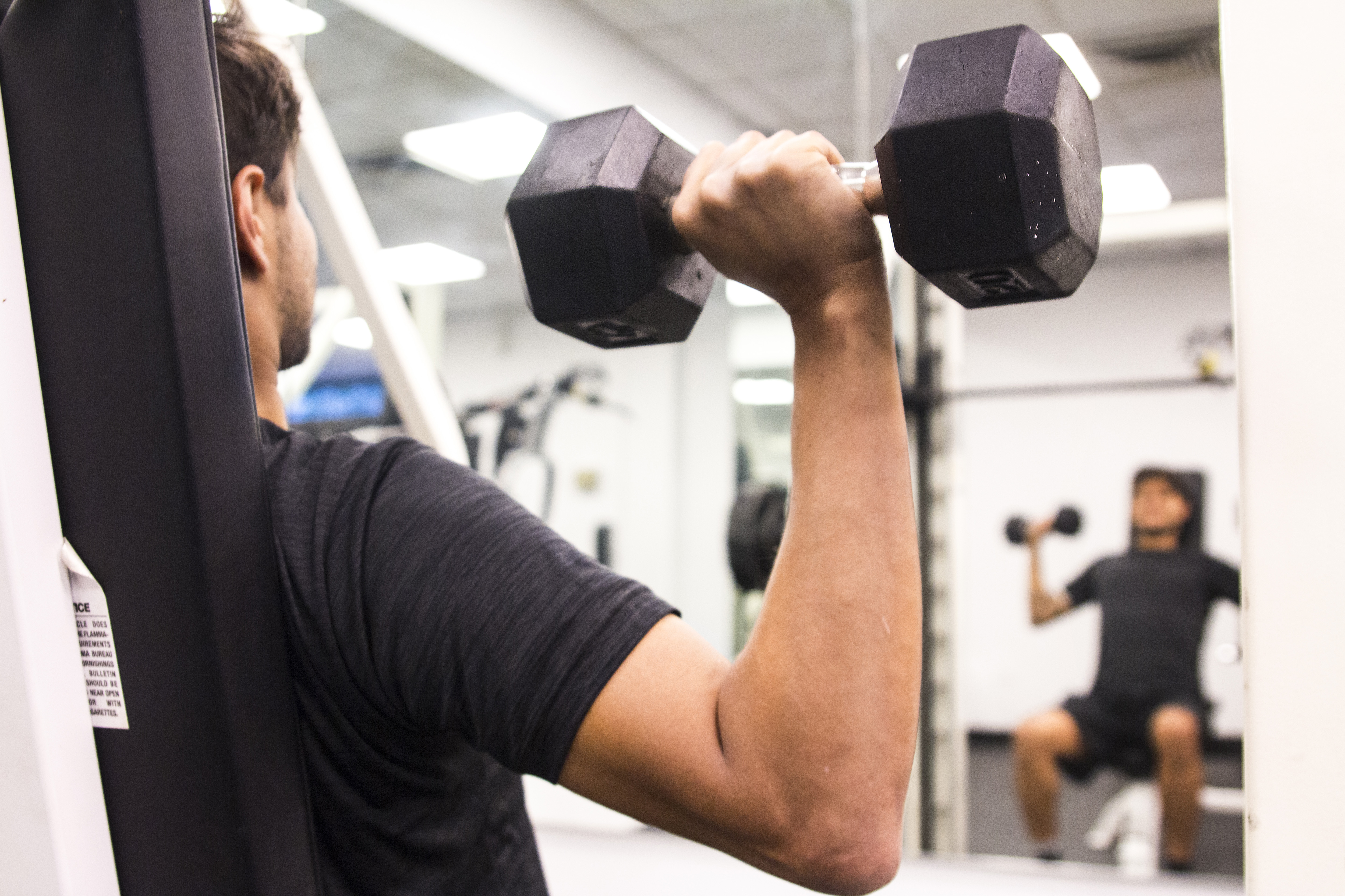 En person sitter på en bänk i ett gym och lyfter en hantel.