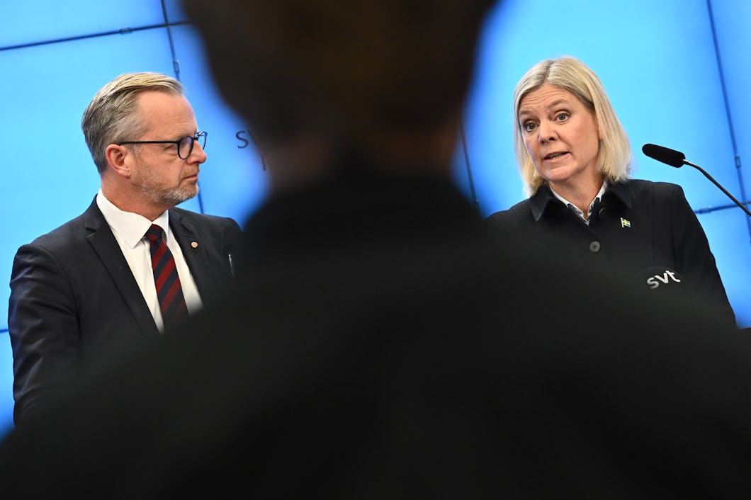 Mikael Damberg i kostym och Magdalena Andersson med blont hår talar på en presskonferens. Bilden är tagen bakom en person som är ur fokus.