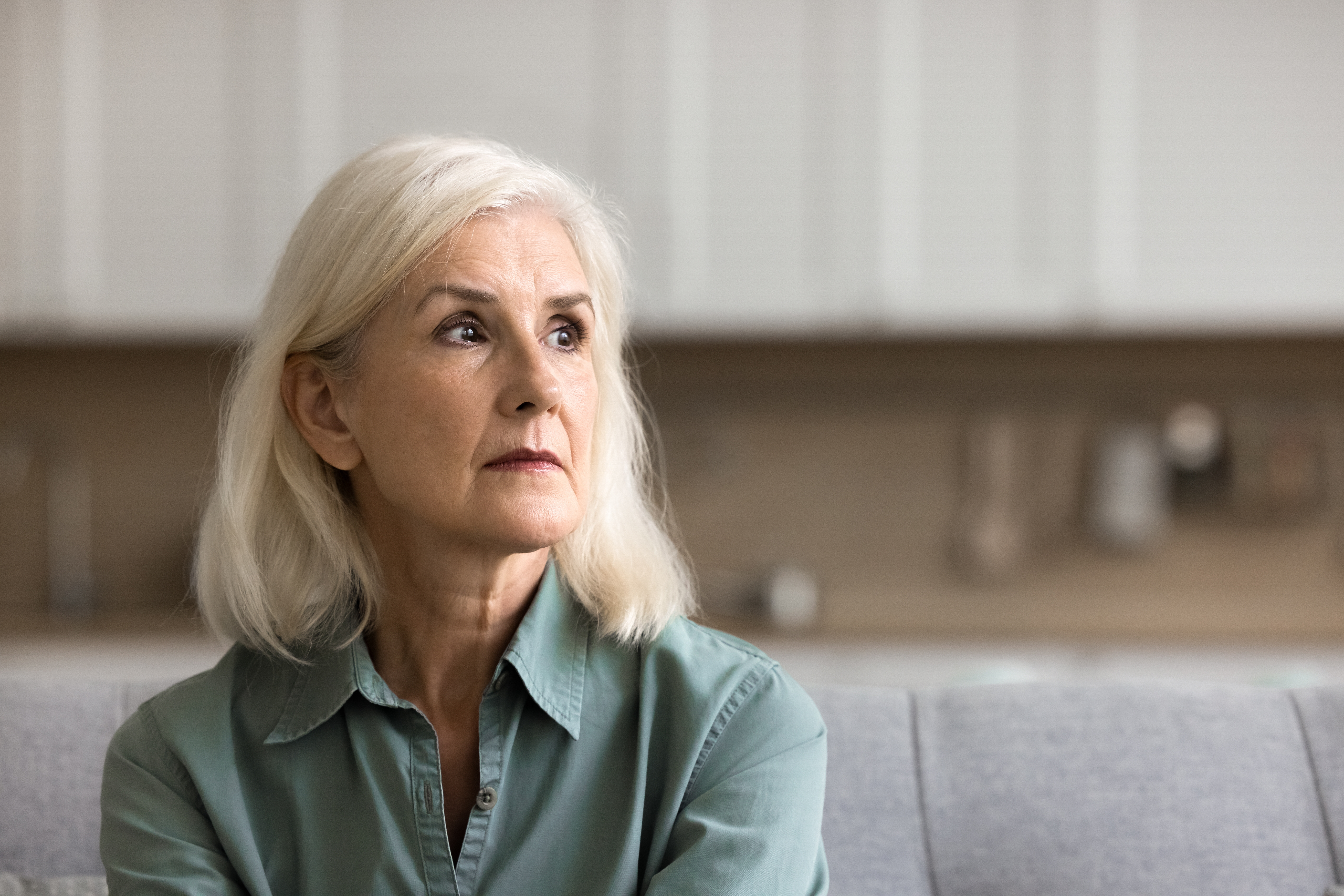 En äldre gråhårig kvinna sitter hemma i en soffa, iklädd grön skjorta och tittar åt sidan.