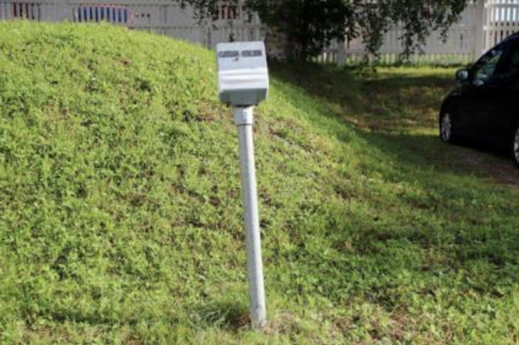 En metallbrevlåda i grå färg är monterad på en stolpe som står på en grässluttning nära en parkerad bil i svart färg.
