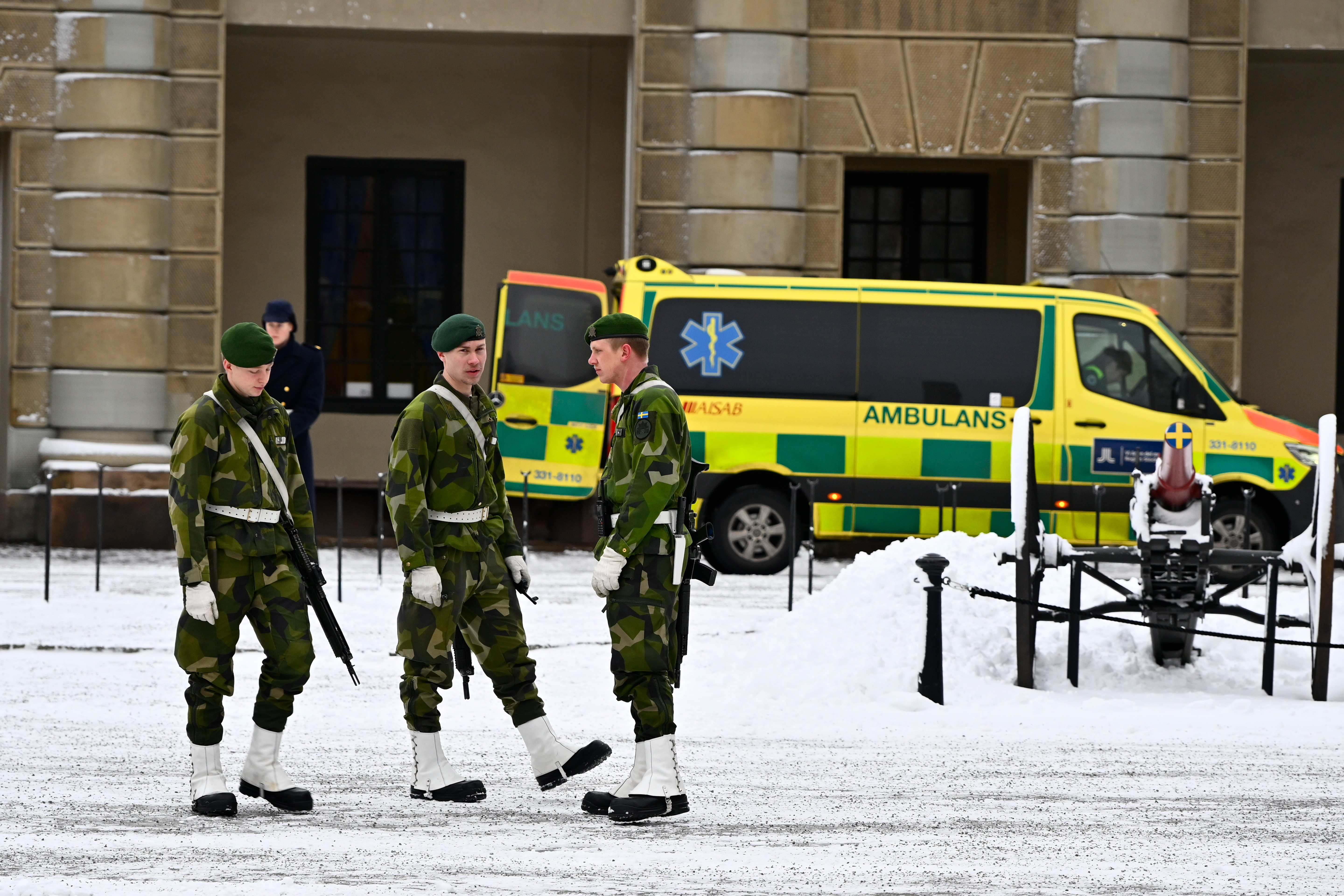 Tre soldater i uniform står på en snötäckt gata intill en gul ambulans. En person går i bakgrunden.
