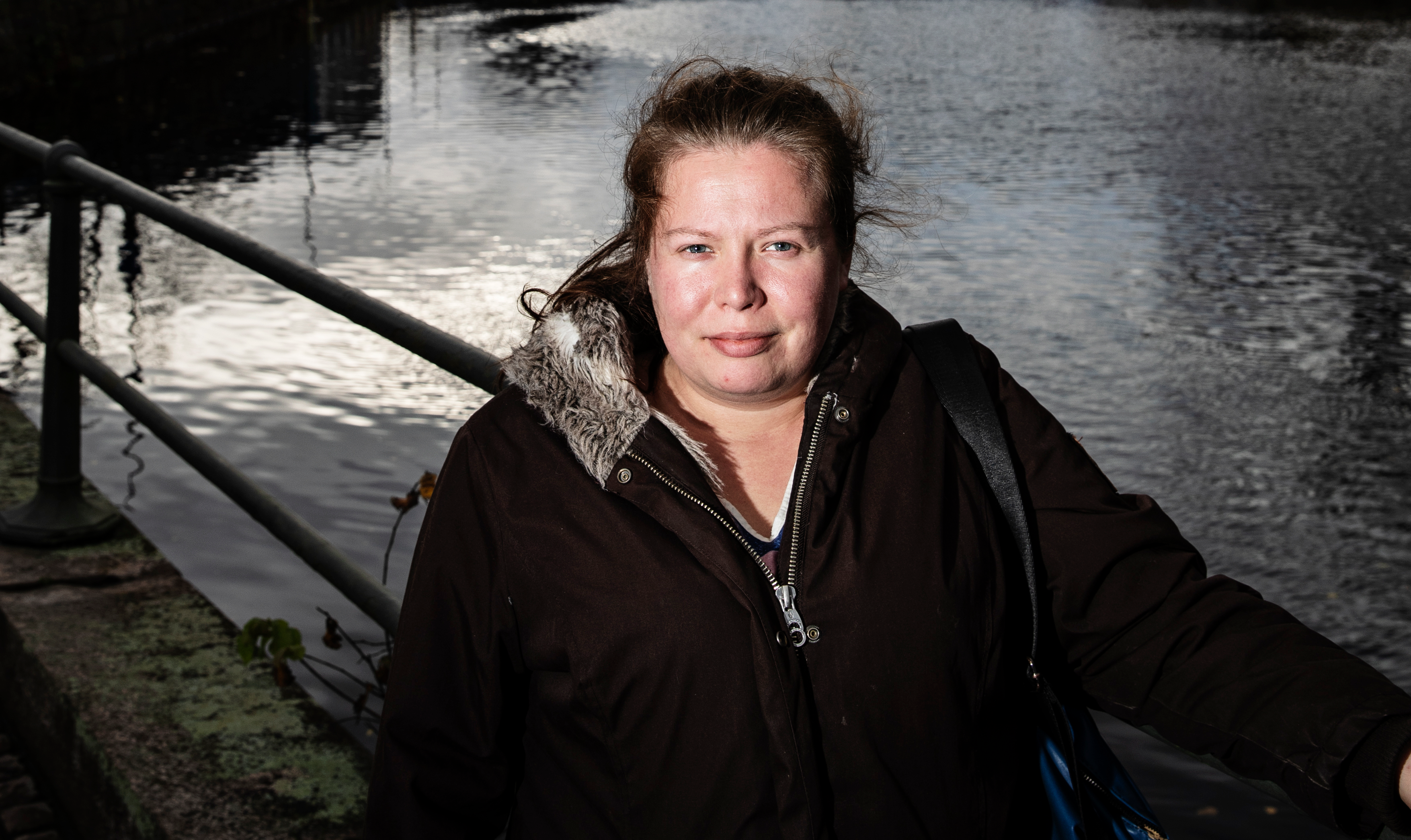 Emelie Edstrom i mörk kappa står vid ett vattendrag med ett metallräcke. I bakgrunden syns vattenytan.