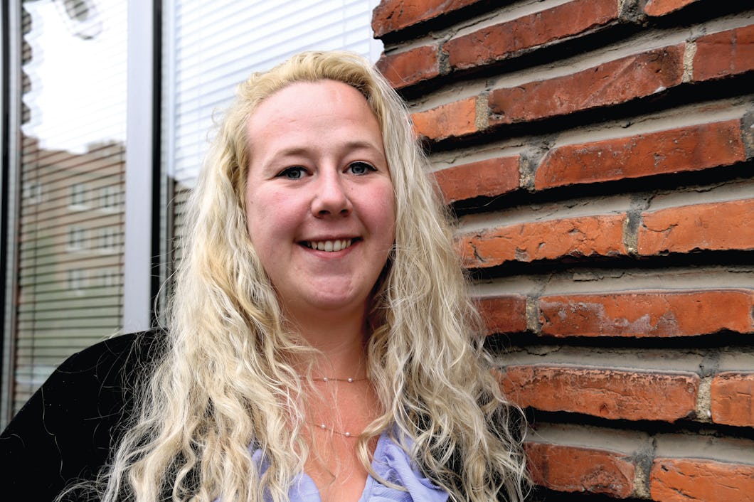 Person med långt blont hår som ler, står bredvid en tegelvägg.