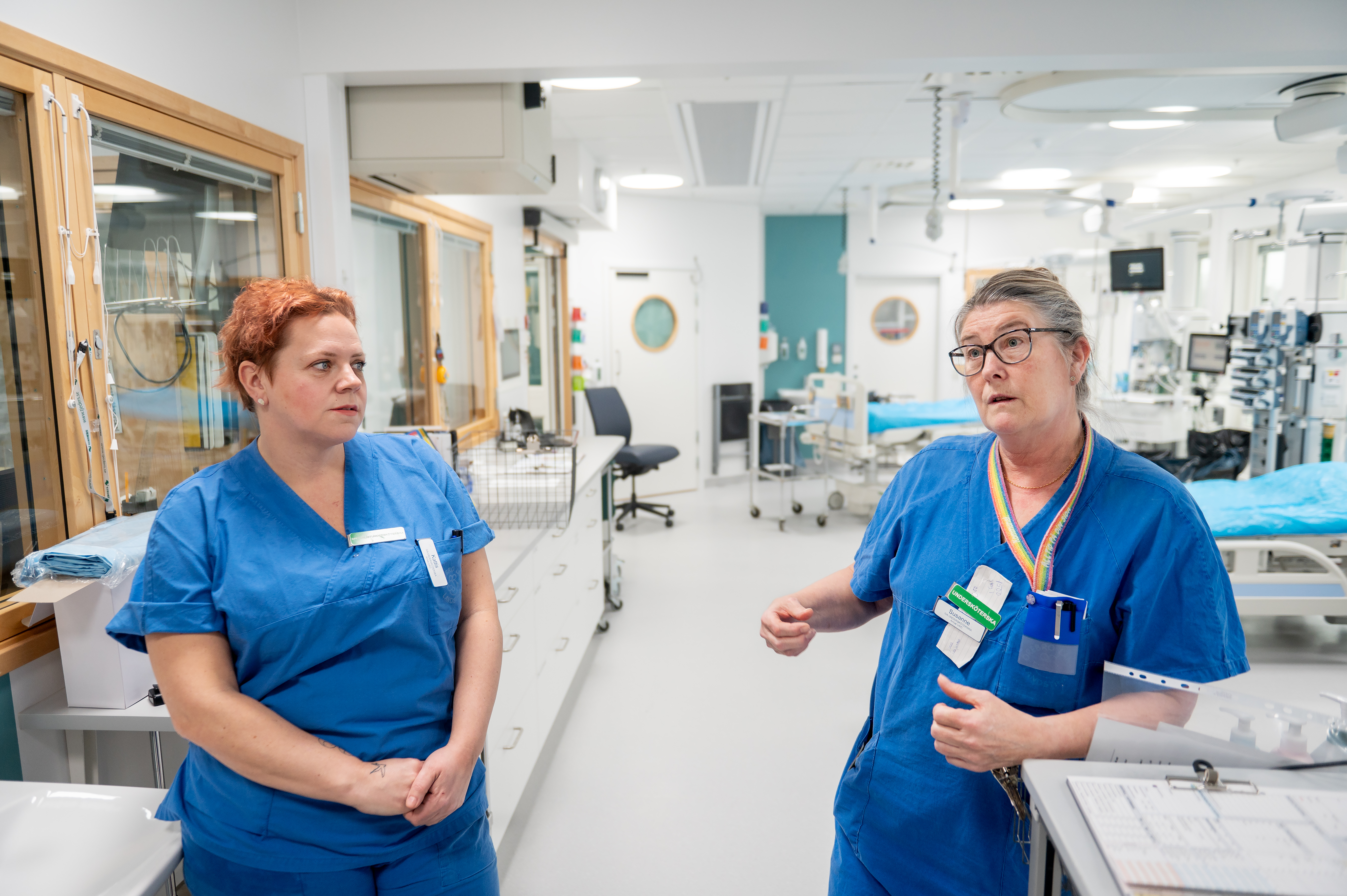 Två vårdpersonal i blå kläder samtalar i en medicinsk anläggning med sängar och medicinsk utrustning i bakgrunden.