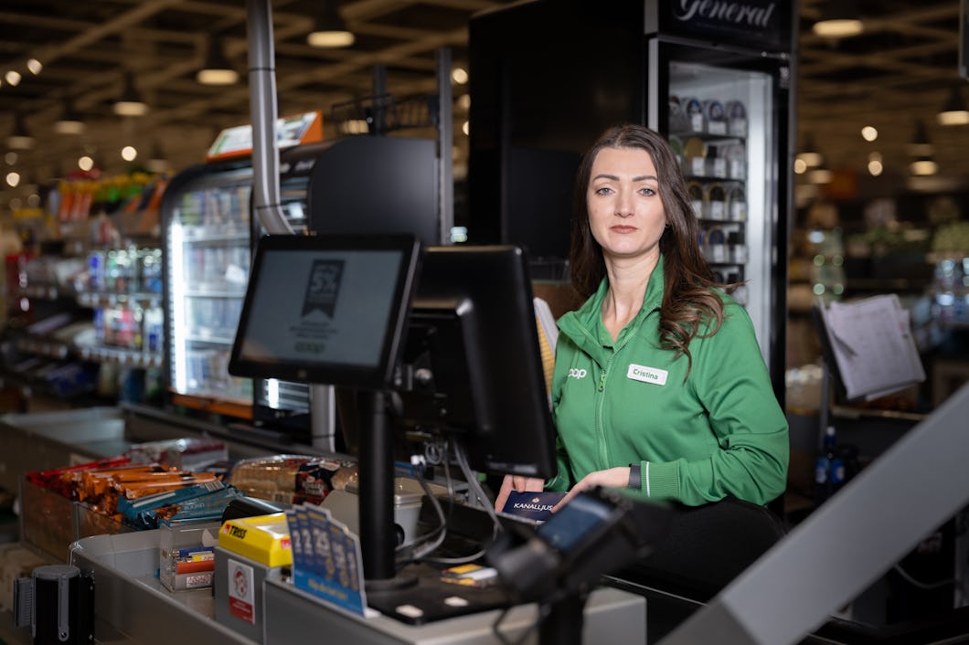 En kassör i grön uniform står vid en kassa i en matbutik, omgiven av snacks och drycker, vänd mot kameran.