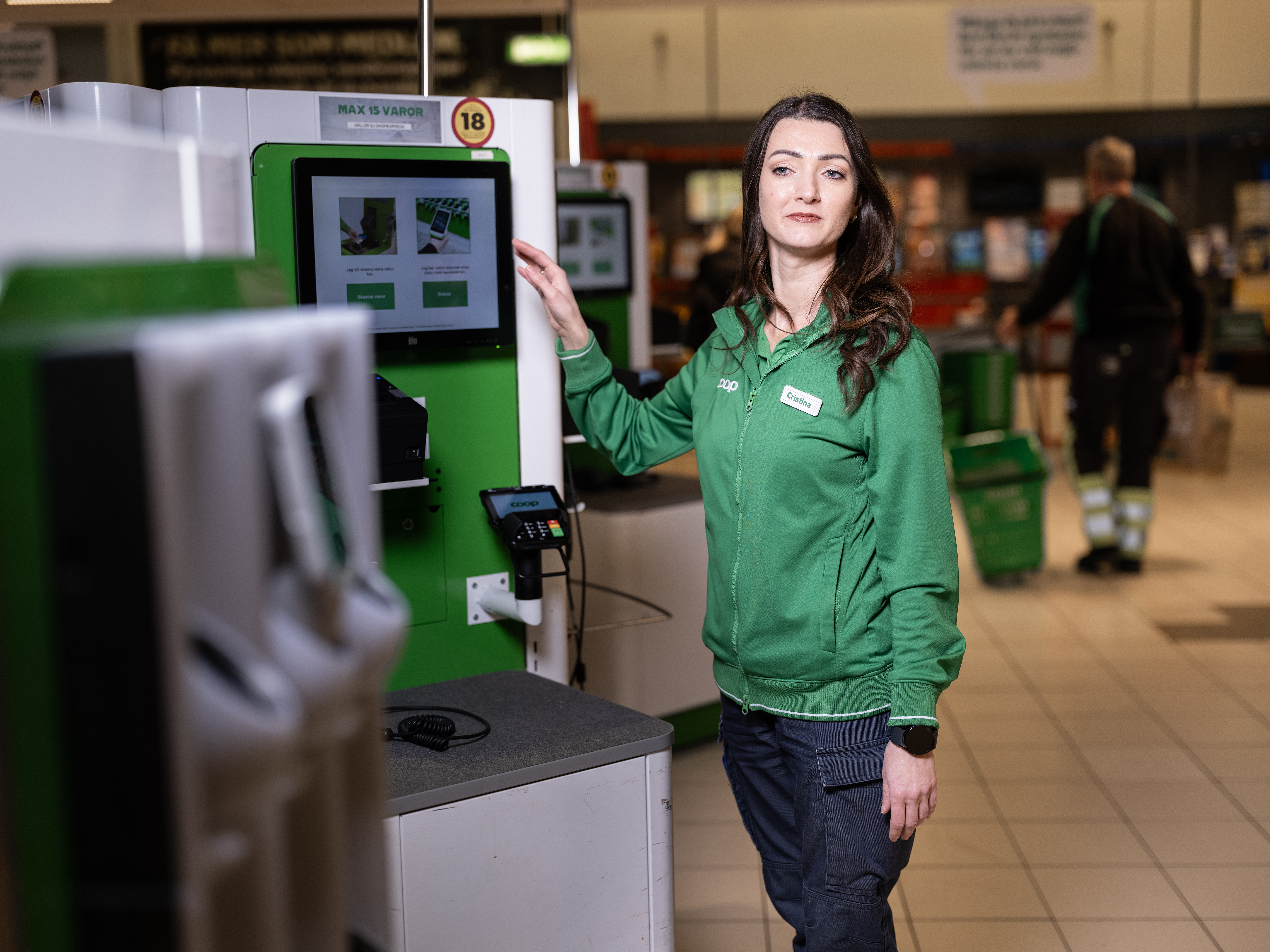 En kvinna i grön jacka står vid en självbetjäningskassa i en butik och tittar mot kameran.