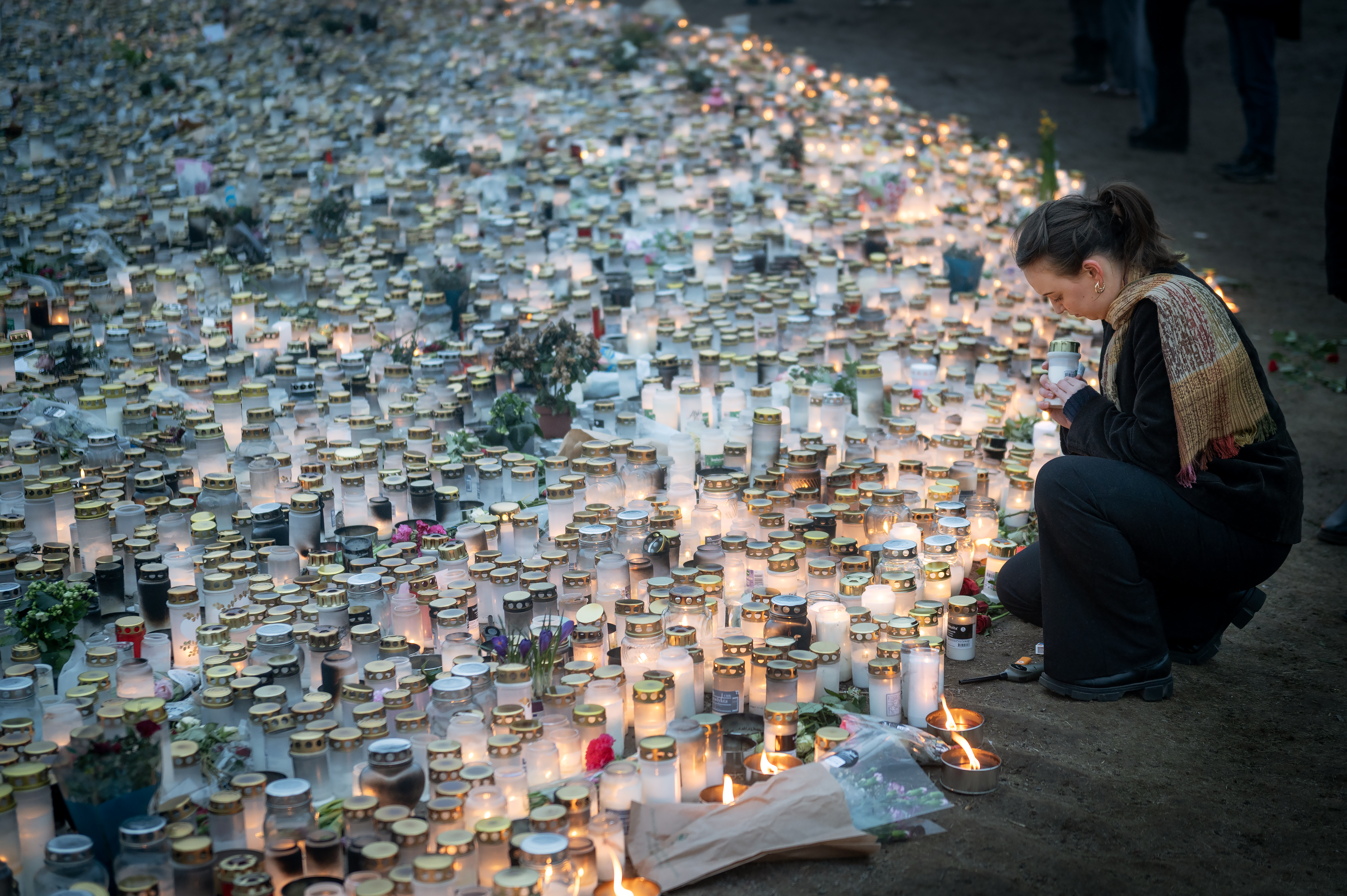 En person knäböjer bland hundratals tända ljus och små burkar, som tillsammans bildar en minnesscen på marken.