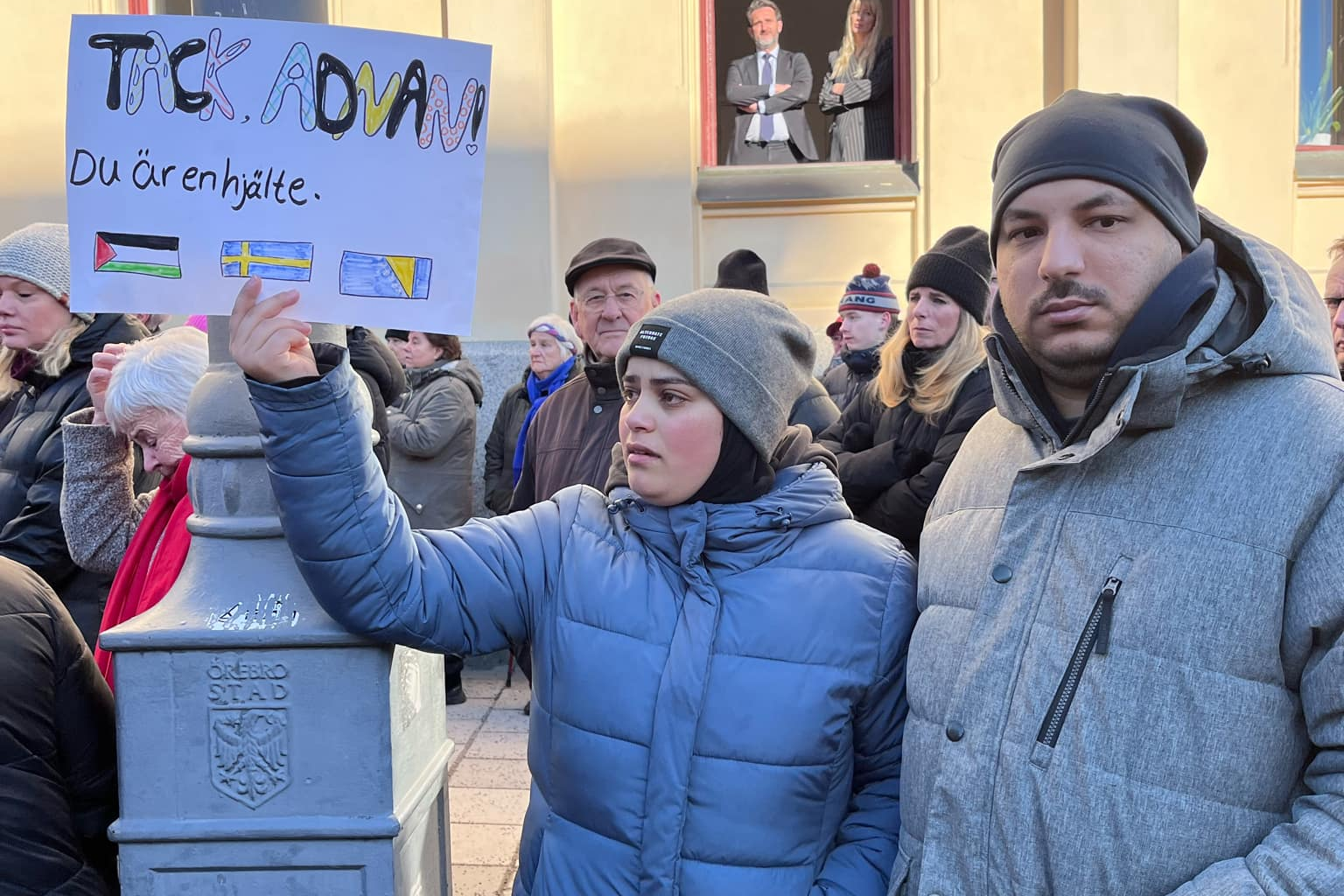 En grupp människor samlade vid en protest. En person håller upp en skylt på svenska samt flaggor. Två individer tittar ner från ett fönster ovanför.