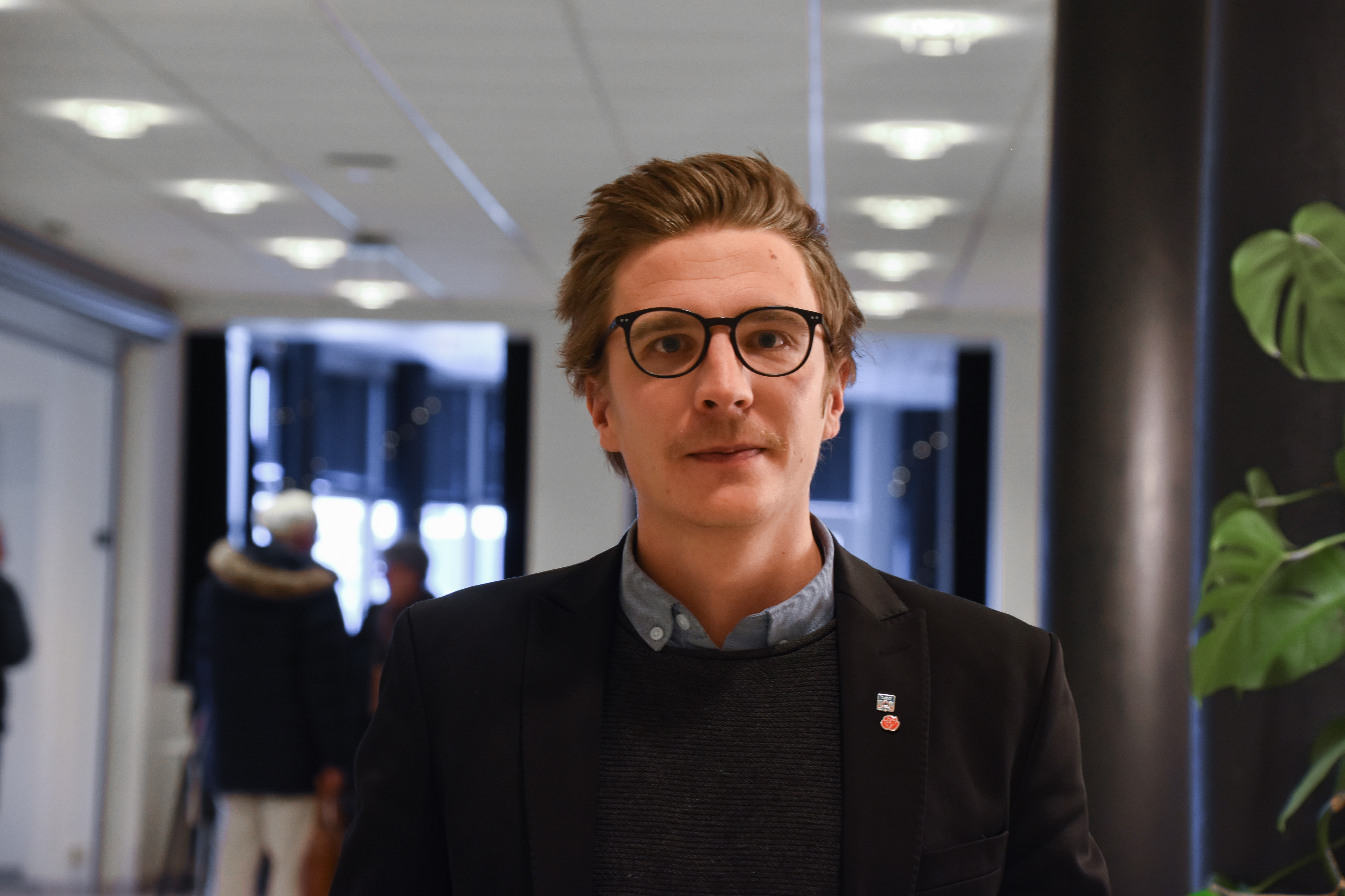 En person med glasögon och kostym står i en inomhusmiljö med otydliga människor i bakgrunden.