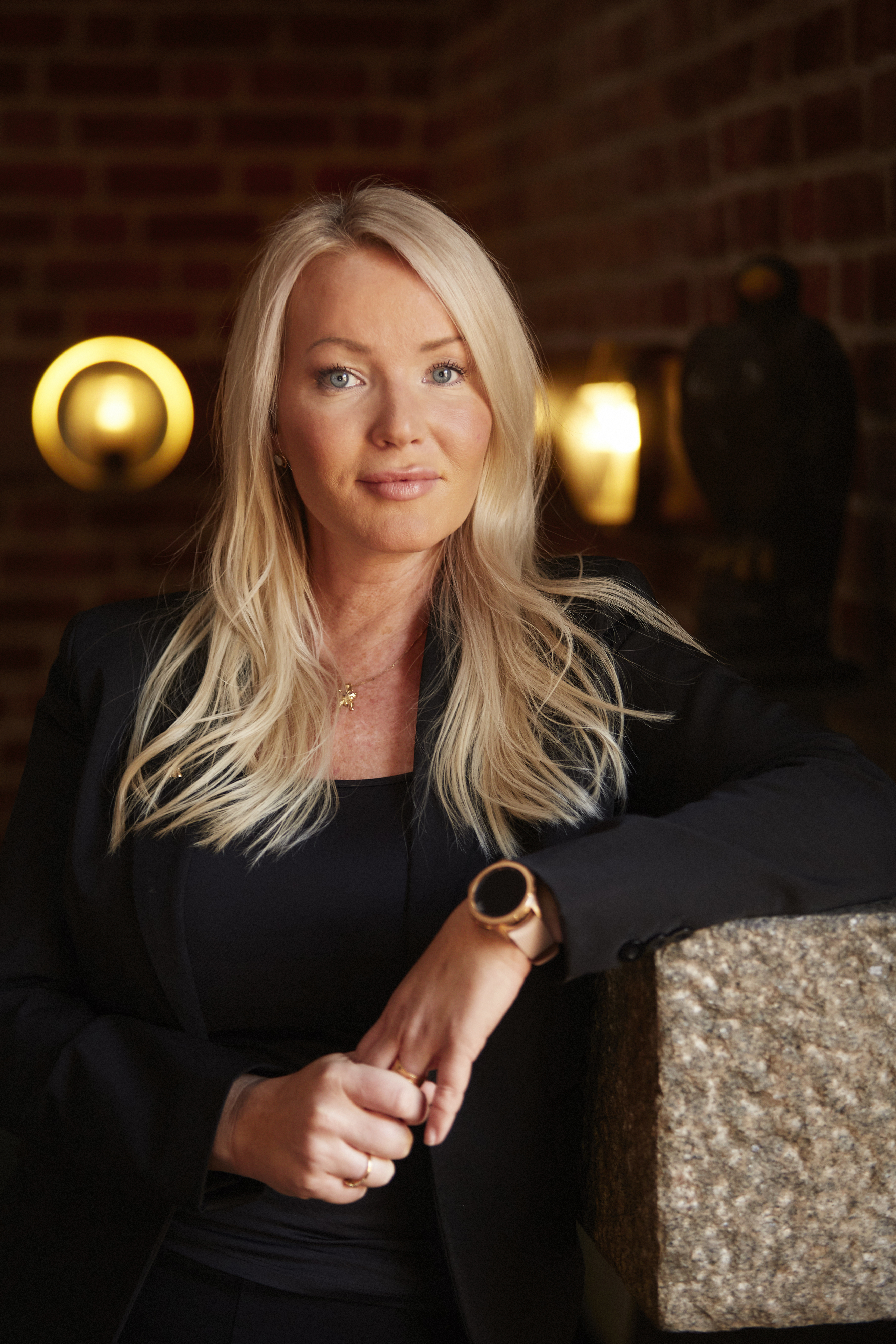 En kvinna med långt blont hår, klädd i en svart kavaj, lutar sig mot en stenliknande yta. Bakgrunden består av en vägg med textur och ljusarmaturer som ger ifrån sig ett varmt sken.