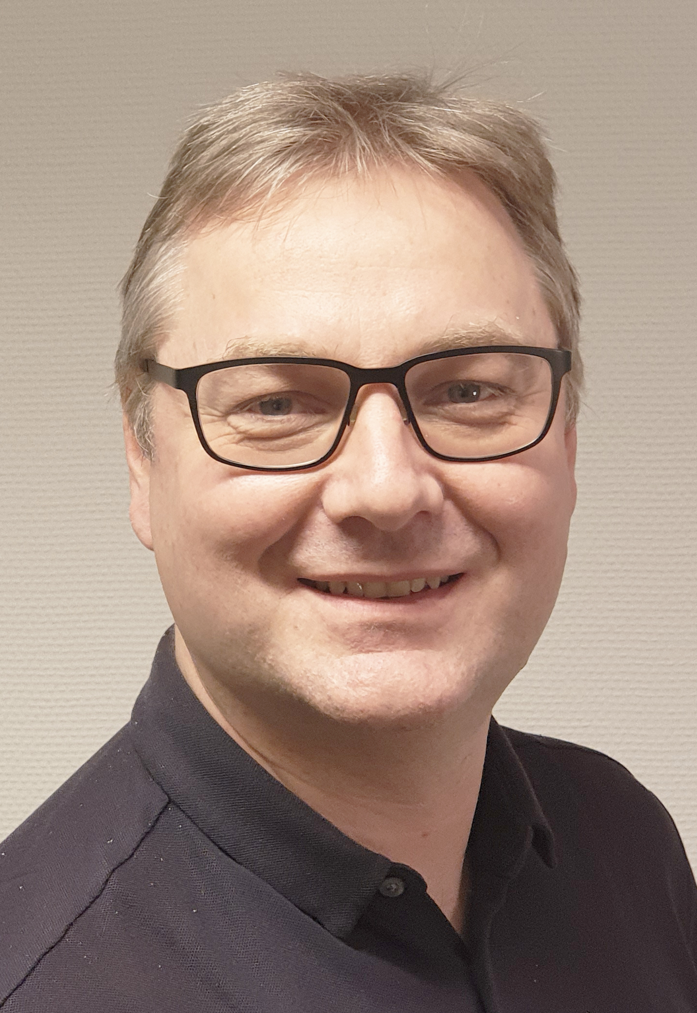 Daniel Modée med kort hår och glasögon, klädd i en mörk skjorta, ler mot kameran framför en ljus bakgrund.