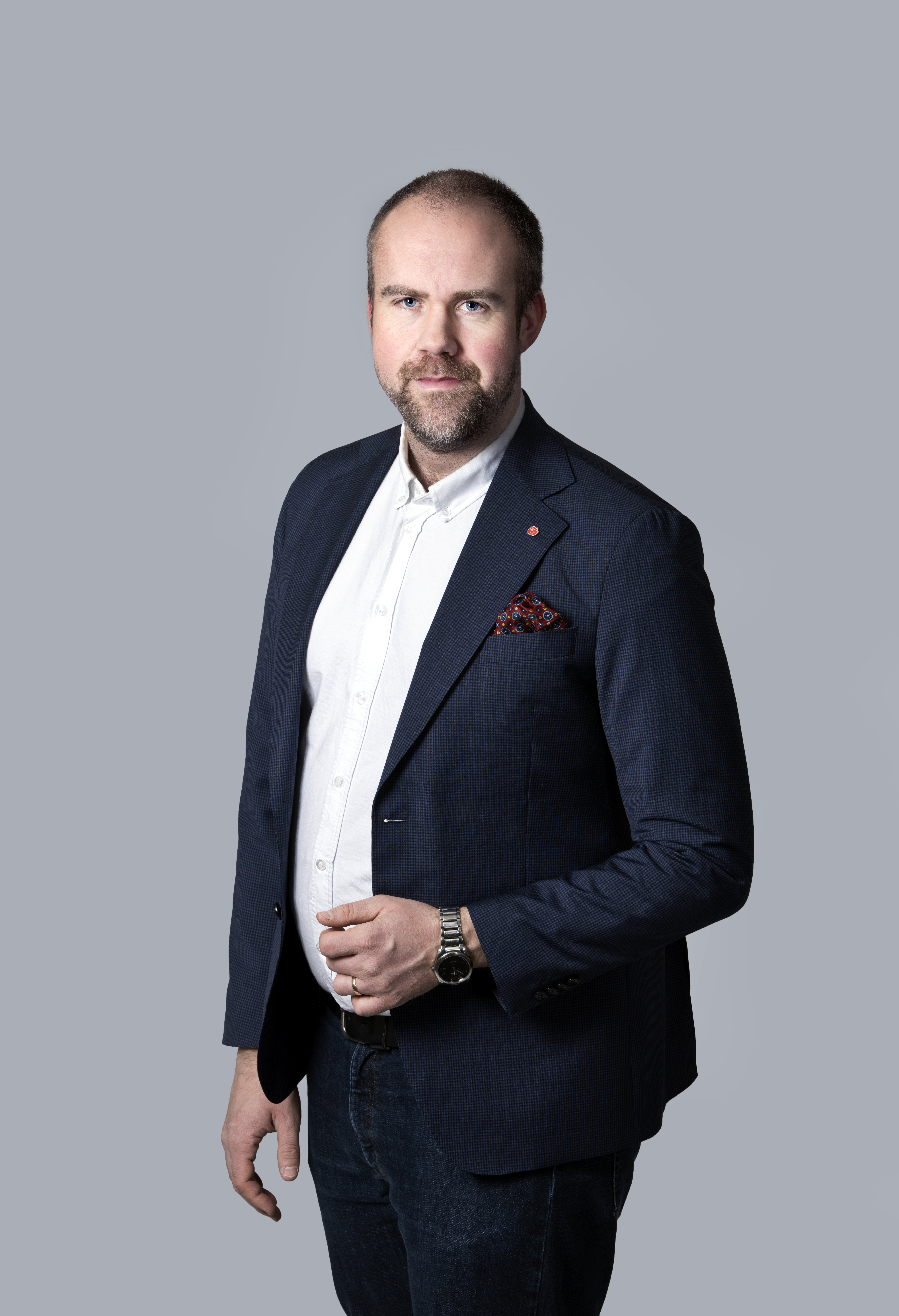 En skäggig man står mot en grå bakgrund, klädd i en mörk kavaj och vit skjorta, med en klocka på handen och ena handen i fickan.