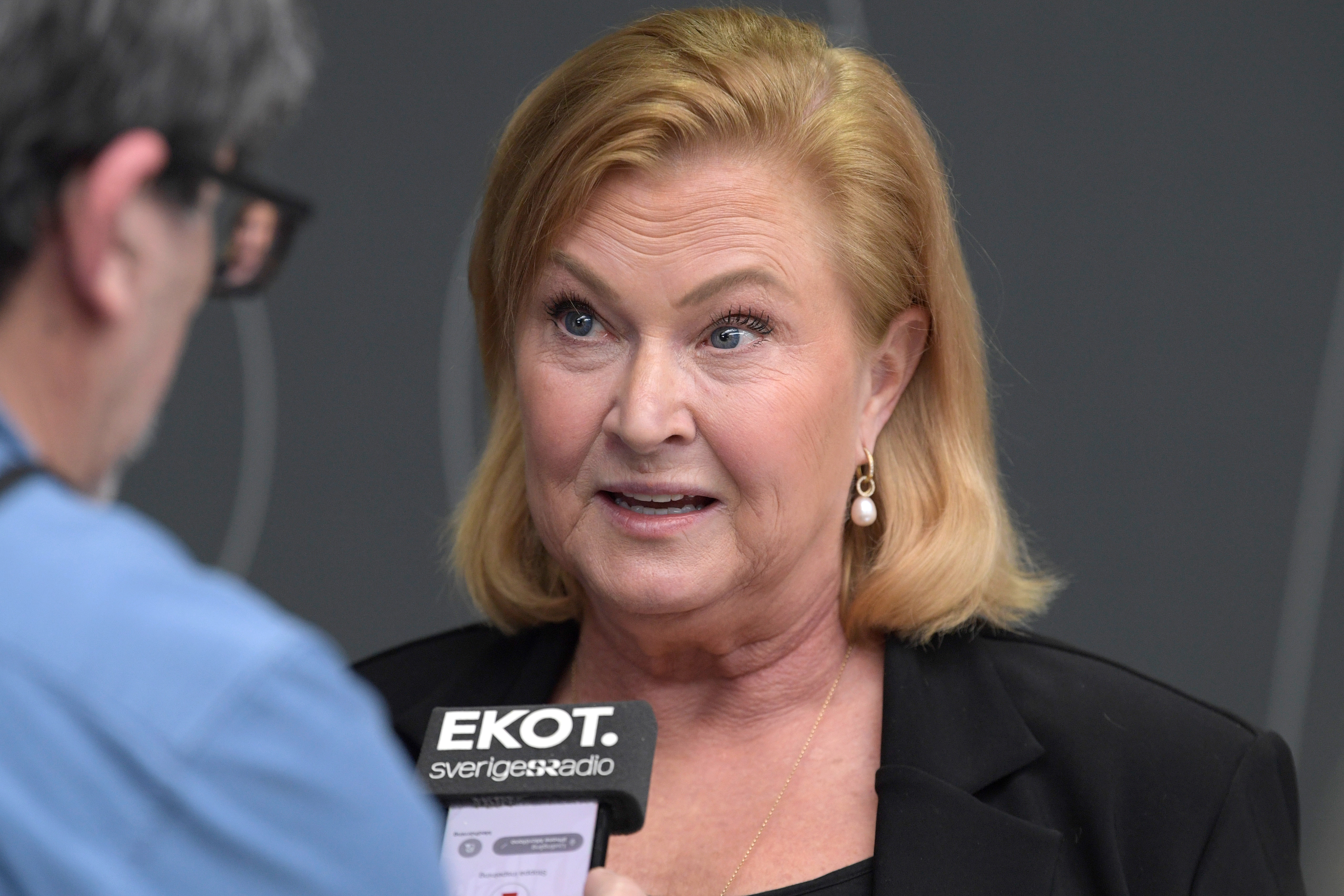 En kvinna med blont hår talar i en mikrofon märkt "EKOT" under en intervju. Hon har på sig en svart blazer och pärlörhängen.