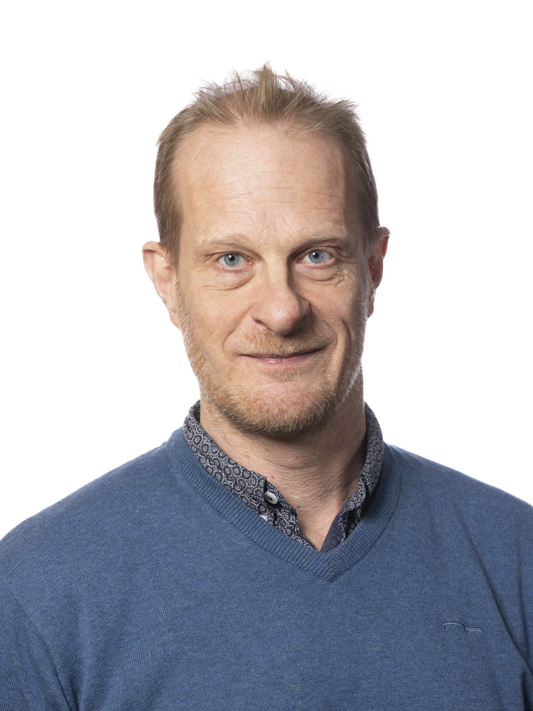 En man i medelåldern med ljust hår och skägg, klädd i en blå tröja och mönstrad skjorta, står mot en vit bakgrund. Hans läpper är något uppåtvända.