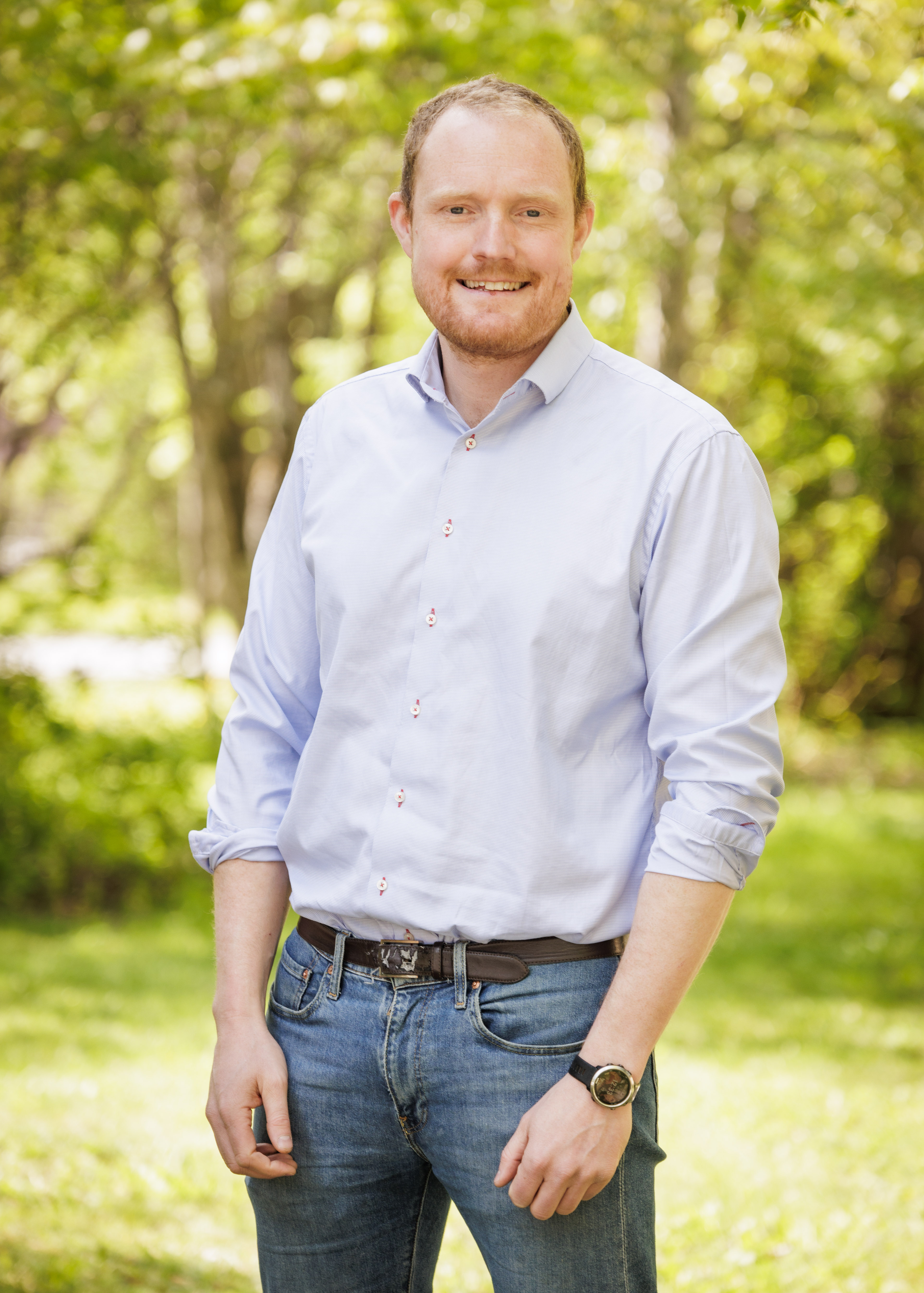 En man i ljusblå skjorta och jeans står utomhus med träd i bakgrunden, och han ler.