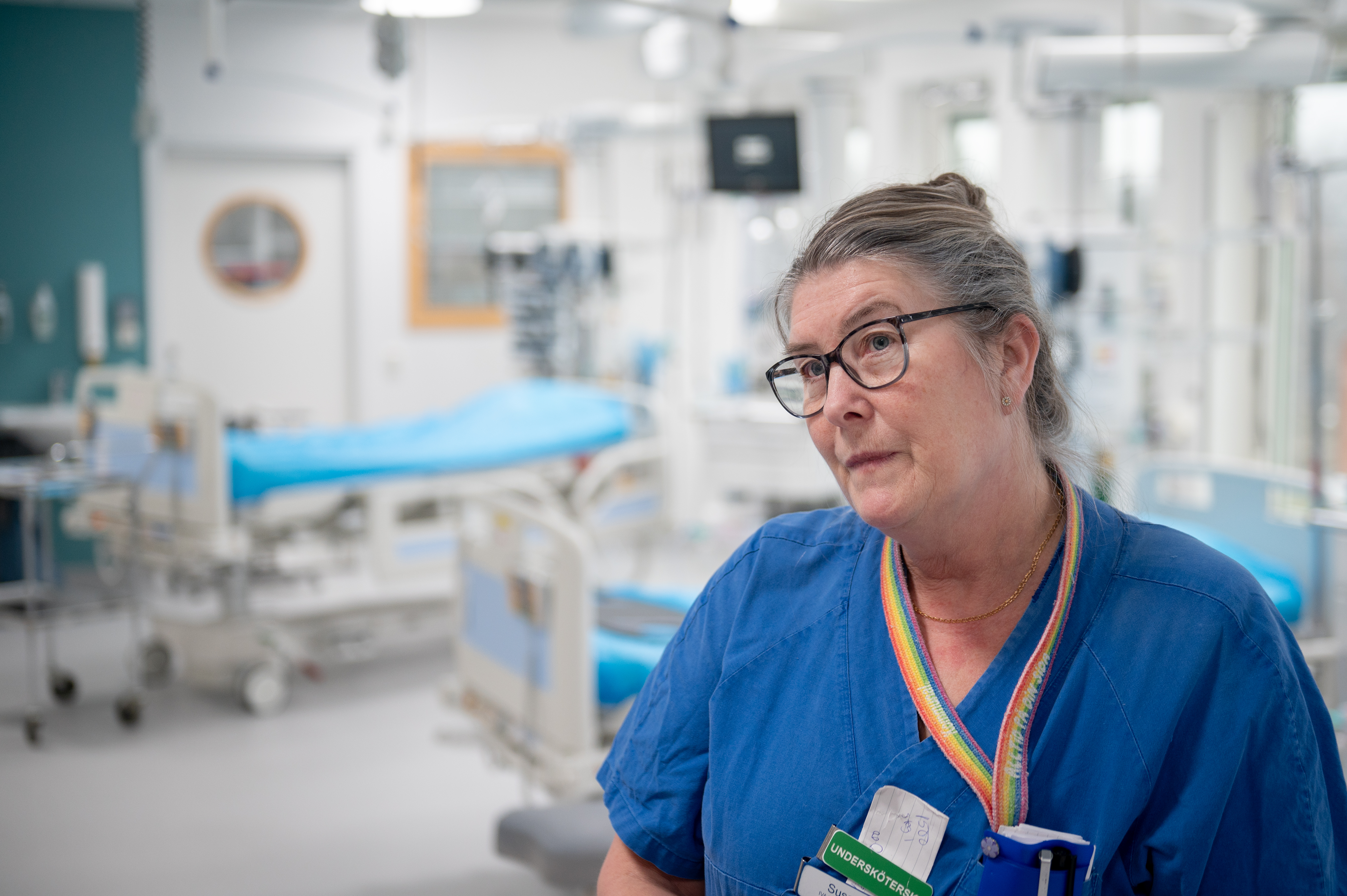 Susanne Jonsson i blå arbetskläder och glasögon står i ett sjukhus med medicinsk utrustning och tomma sängar i bakgrunden.
