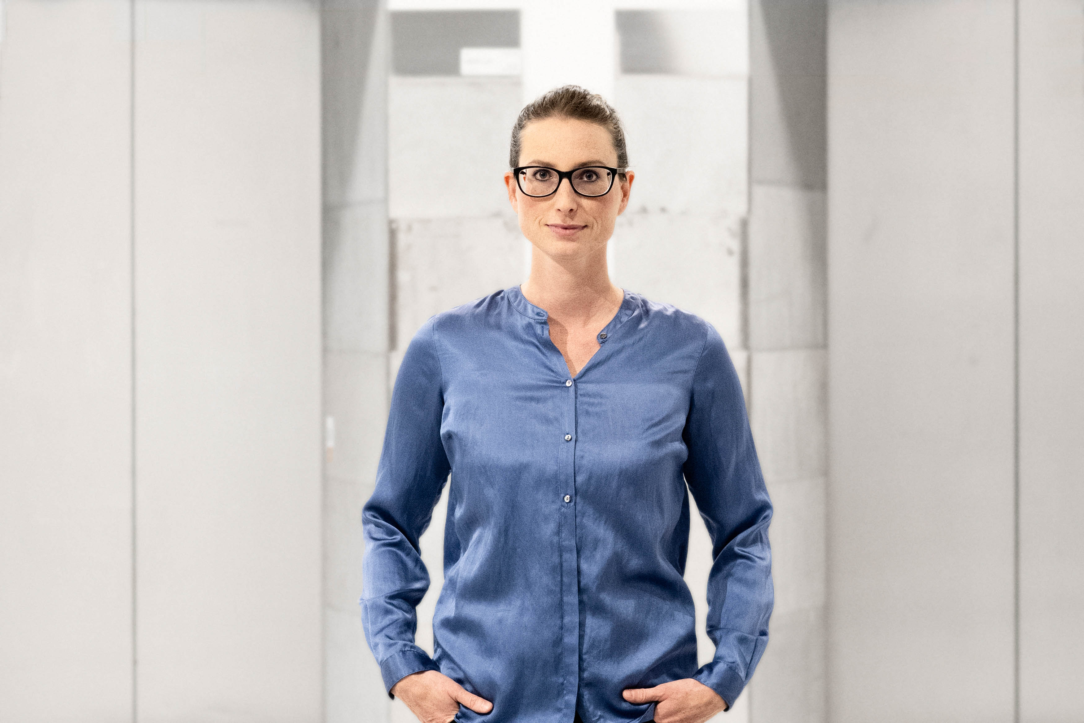 Sofia Viotti med glasögon och en blå skjorta står med händerna i fickorna, omgiven av grå väggar.