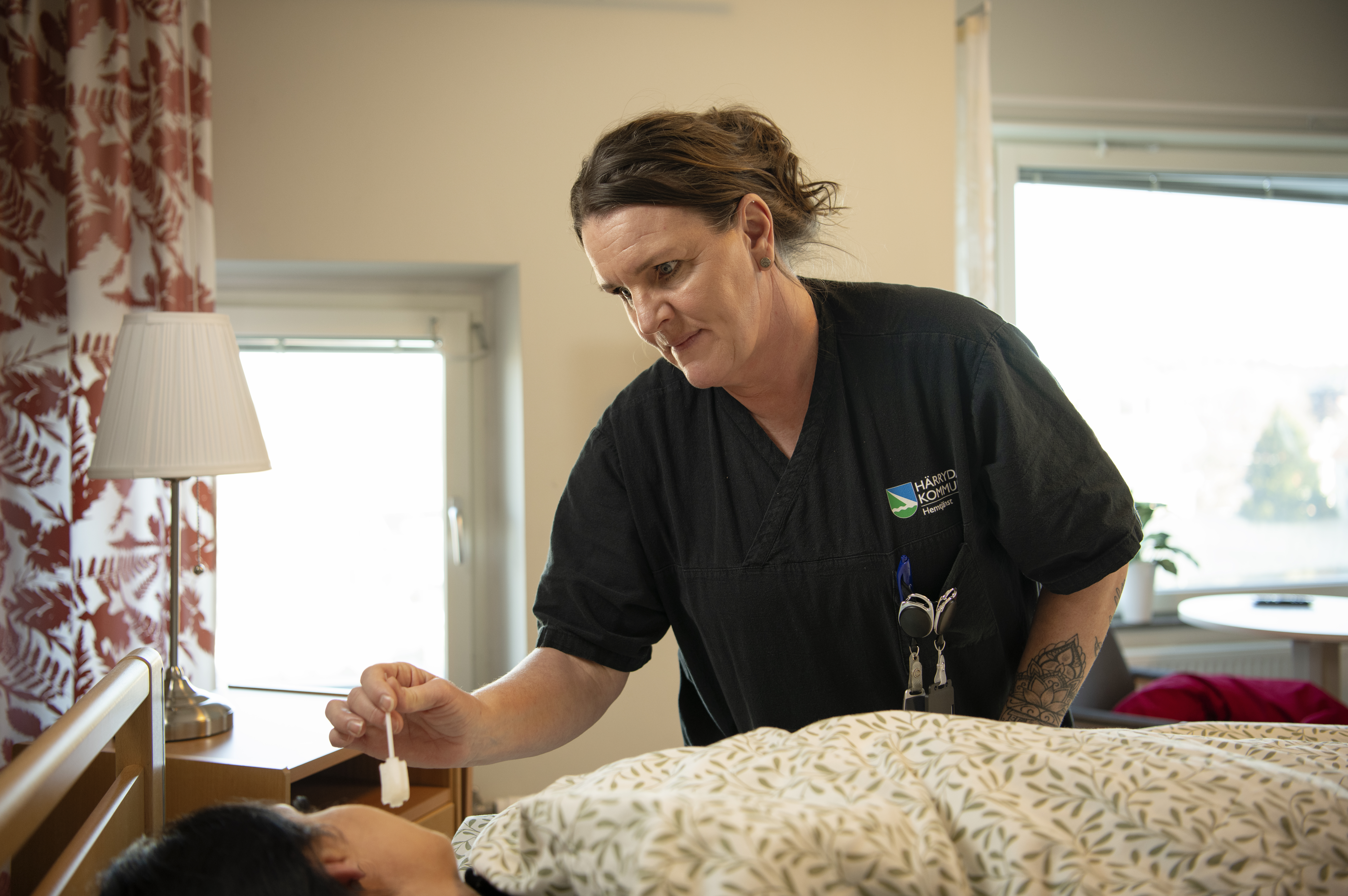 En vårdarbetare. Monica Stenmark, i svart uniform tar hand om en person som ligger i sängen och håller en liten pipett. Rummet har en lampa och ett fönster med gardiner.