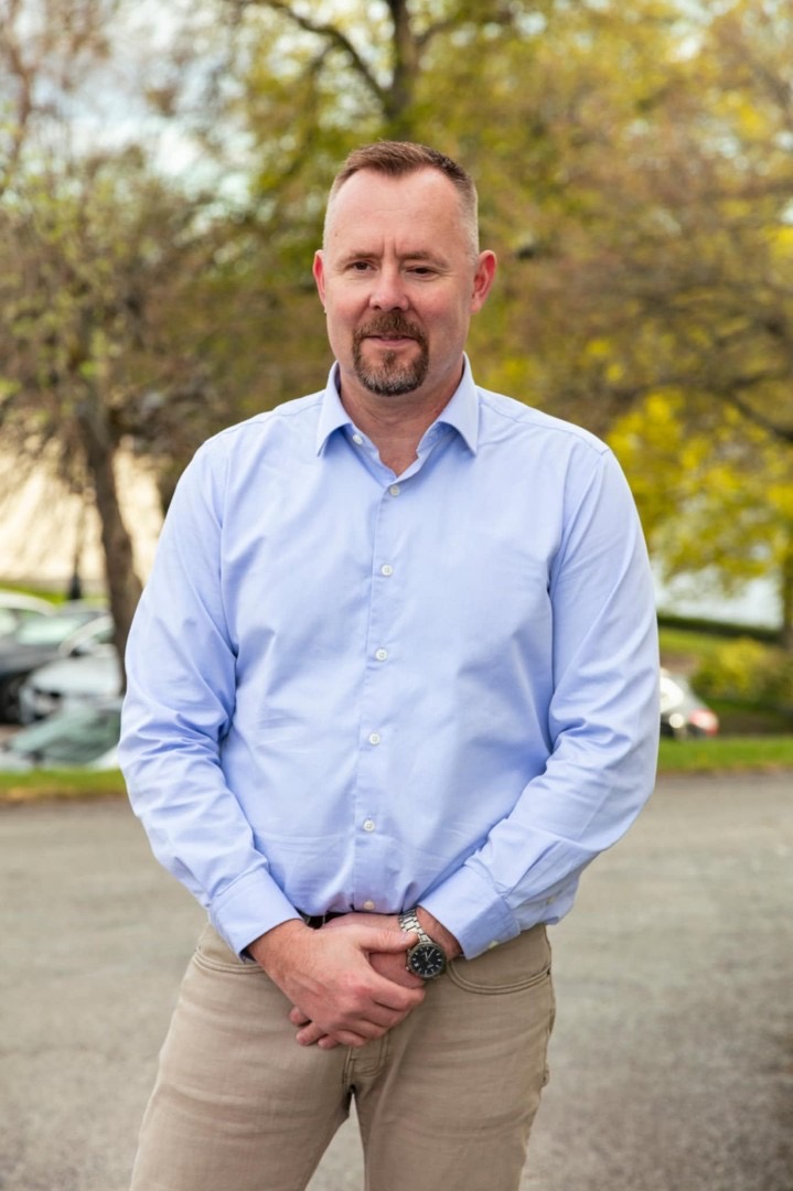 En person med en ljusblå skjorta och beige byxor står på ett asfalterat område. I bakgrunden syns träd och bilar.