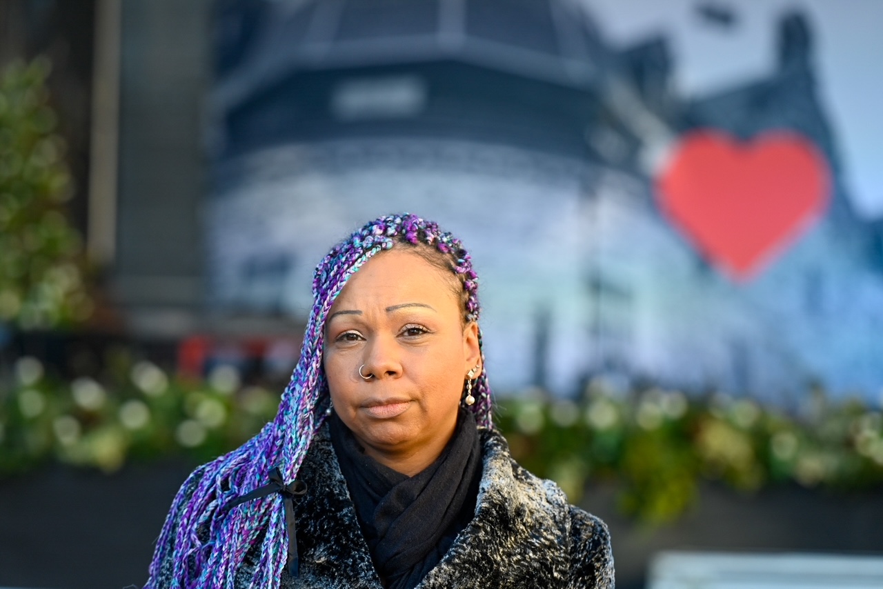 Grace Zuleta med lila och blå flätor står utomhus, iklädd en svart halsduk och en kappa med pälskrage. I bakgrunden syns en suddig väggmålning med ett rött hjärta.