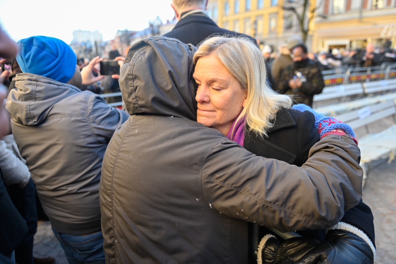 Magdalena Andersson (S) i en kappa kramar någon i en folkmassa utomhus, omgiven av människor som tar fotografier.