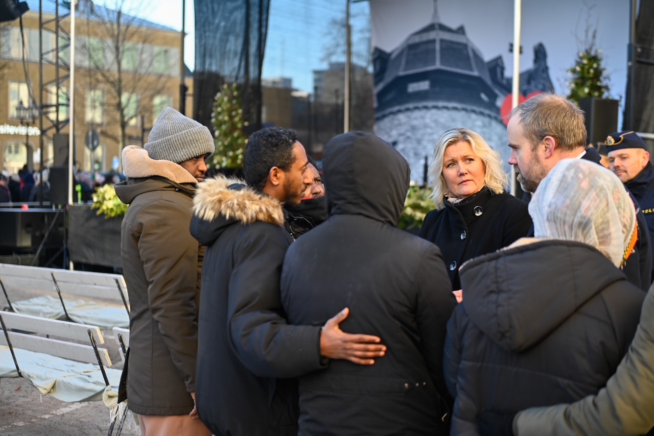 En grupp personer iförda vinterkläder står samlade utomhus och samtal. I bakgrunden syns en banderoll och byggnader.