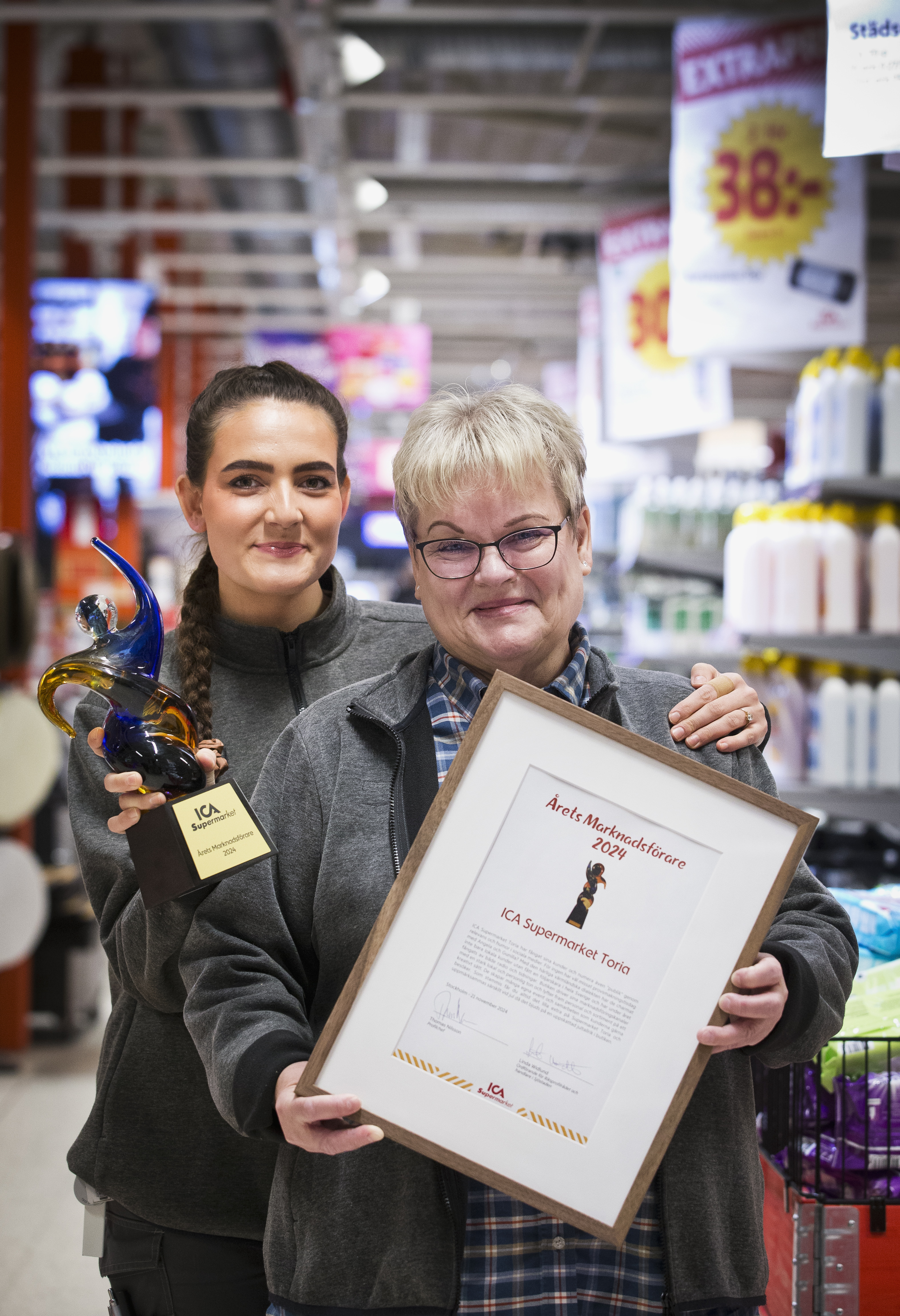 Två personer i en butik håller i en utmärkelse och ett inramat certifikat. I bakgrunden syns hyllor med produkter.