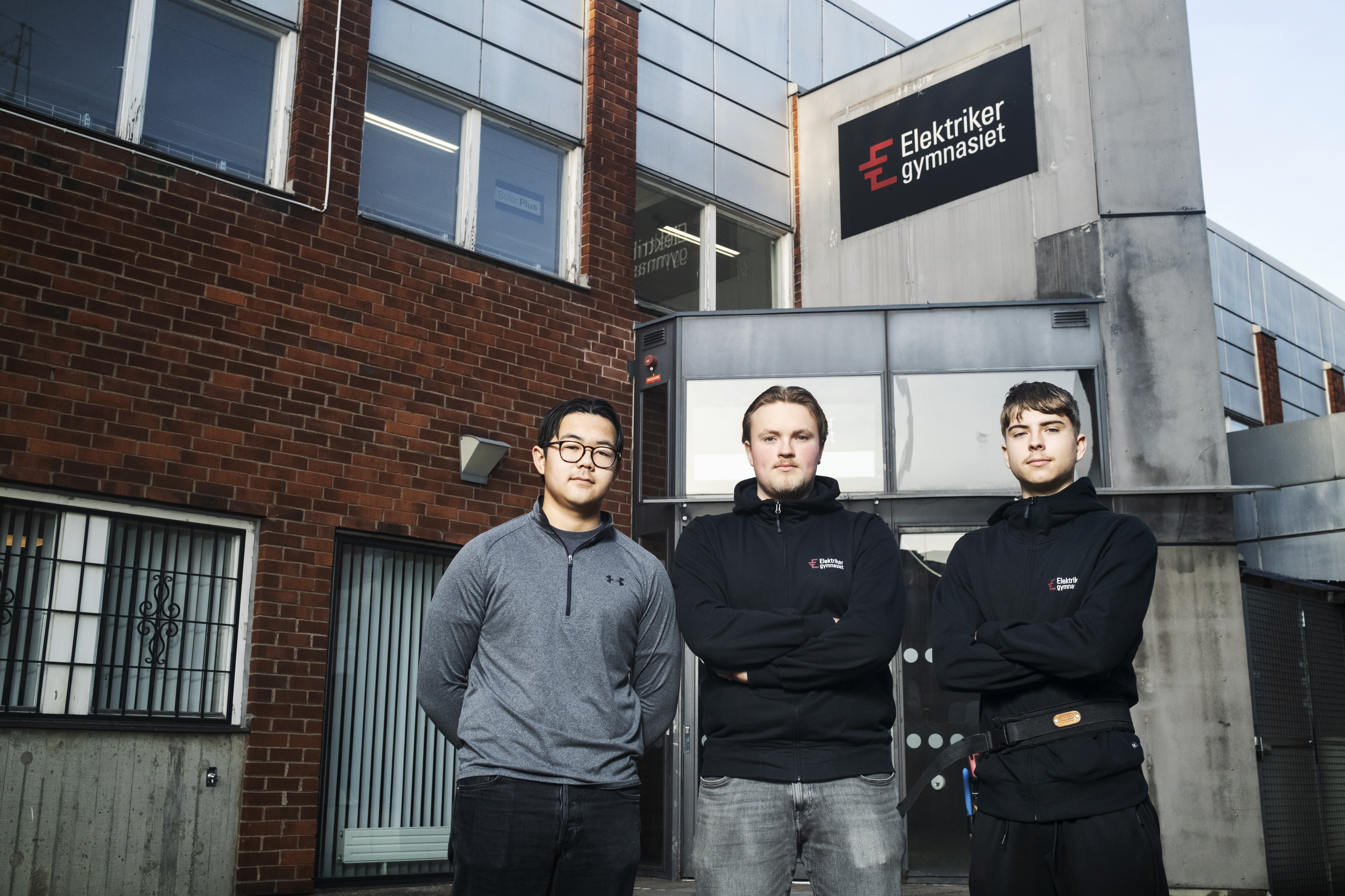 Tre personer står framför Elektriker Gymnasiet, en byggnad med inslag av tegel och glas. De poserar för fotot med armarna i kors.