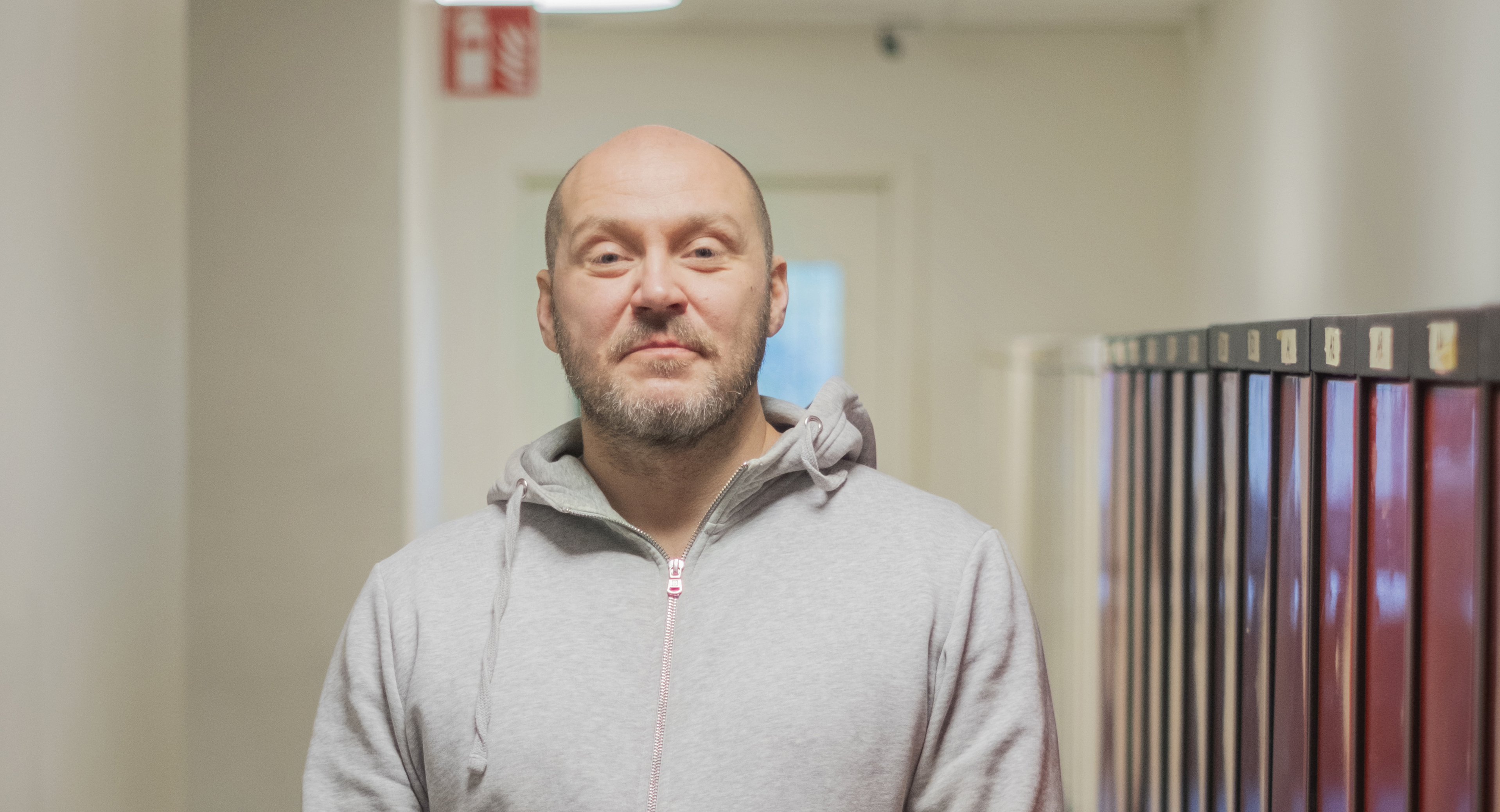 En person i en grå huvtröja står i en korridor bredvid röda skåp, med en brandlarmsskylt synlig i bakgrunden.