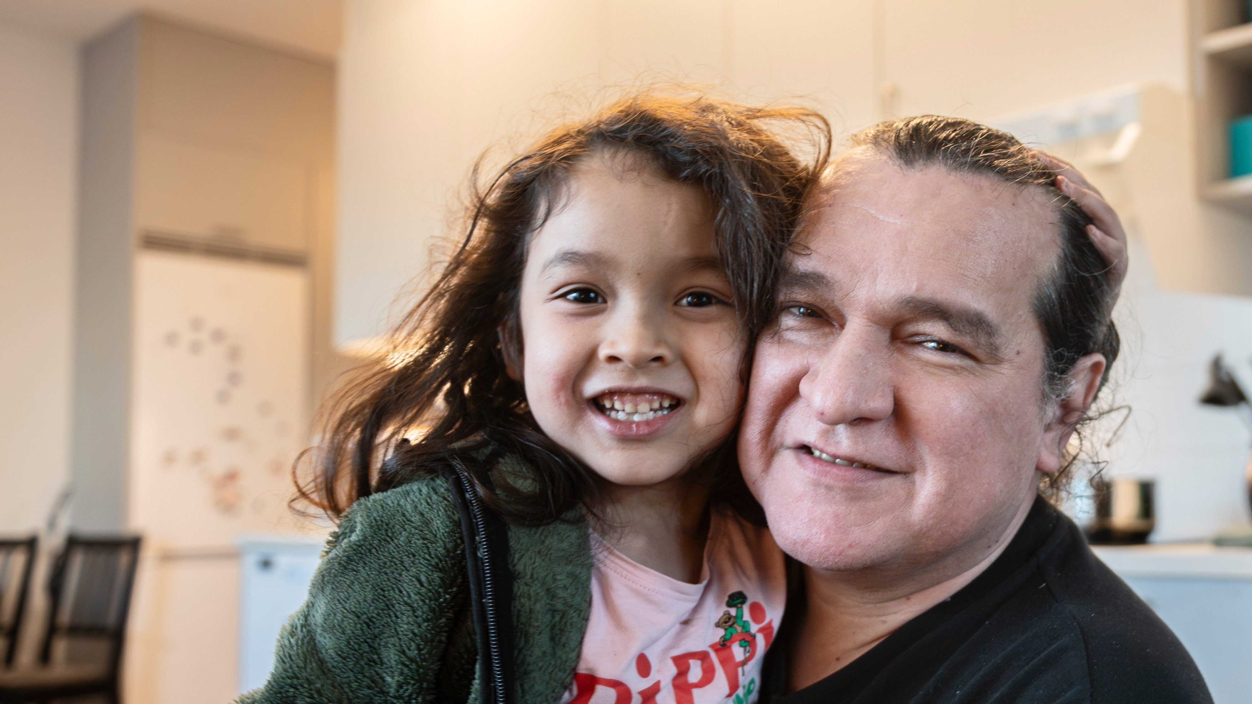 Pavel Ramirez med ett barn som ler i ett rum med varm belysning. Barnet har långt hår och en grön jacka, medan den vuxna har axellångt hår och en svart skjorta. De är nära varandra.