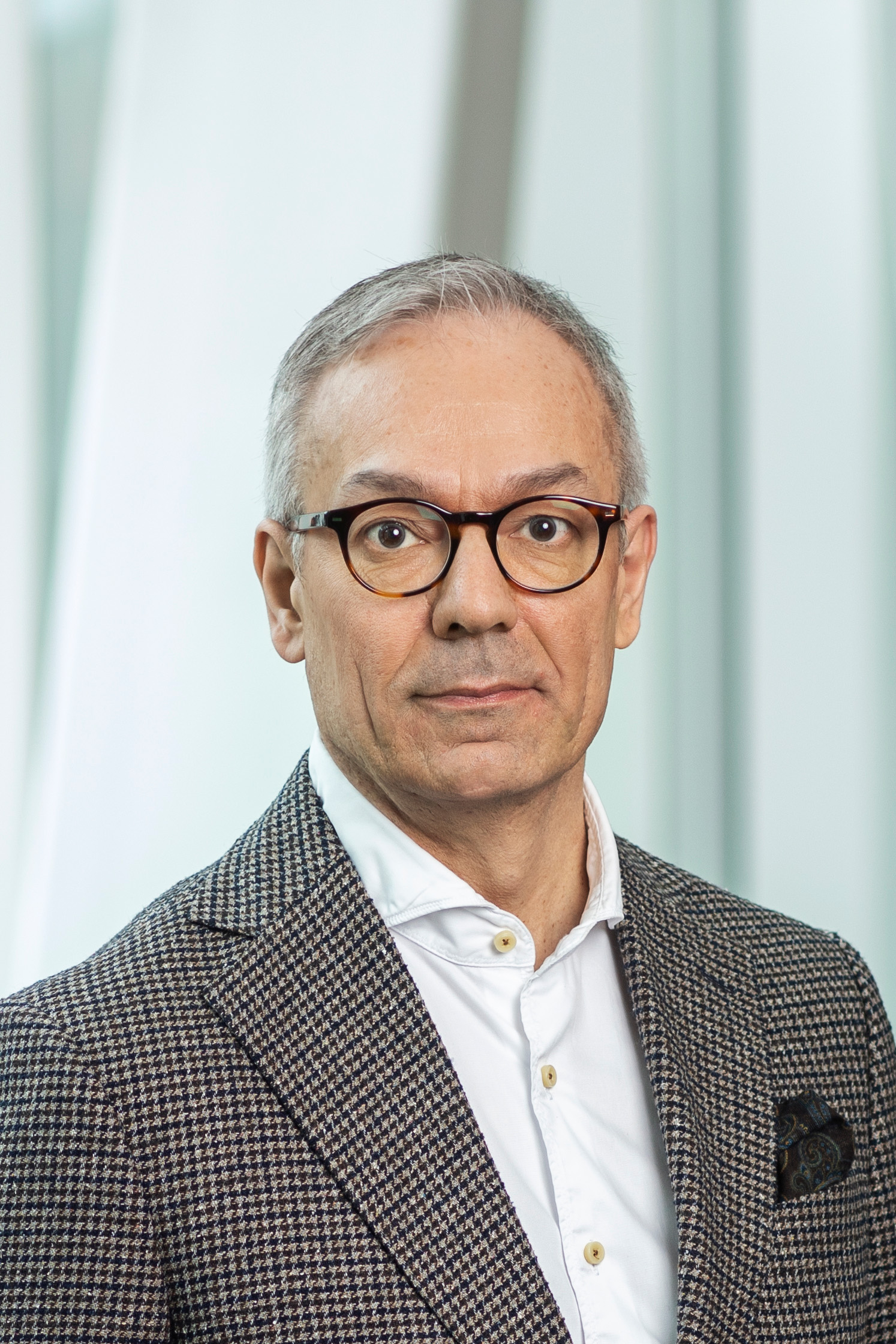 Arturo Arques med kort grått hår och glasögon, iklädd en kavaj med mönster och vit skjorta, står mot en oskarp ljus bakgrund.